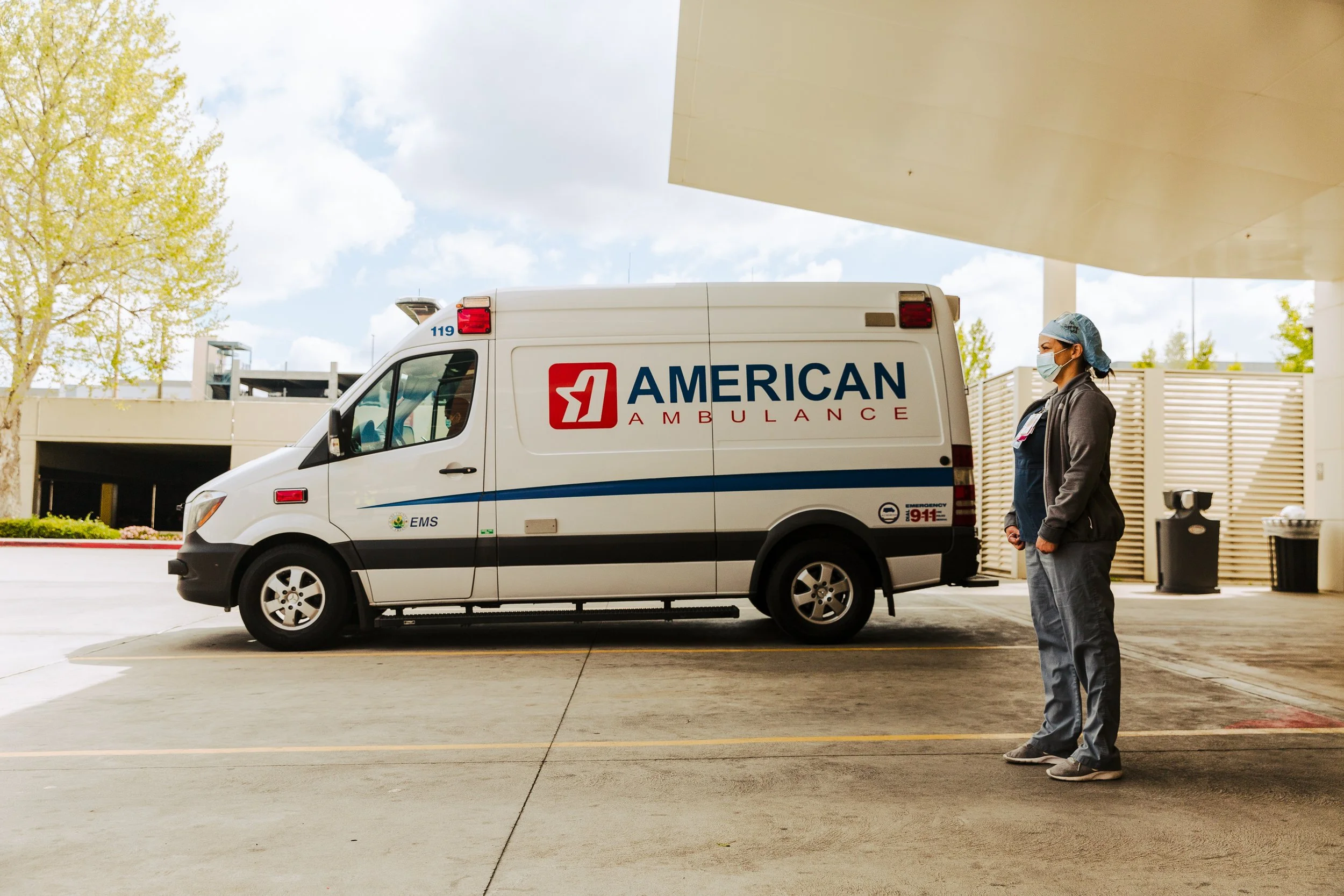 A healthcare worker standing by an ambulance outside a hospital, wearing a face mask, surgical cap, and scrubs.