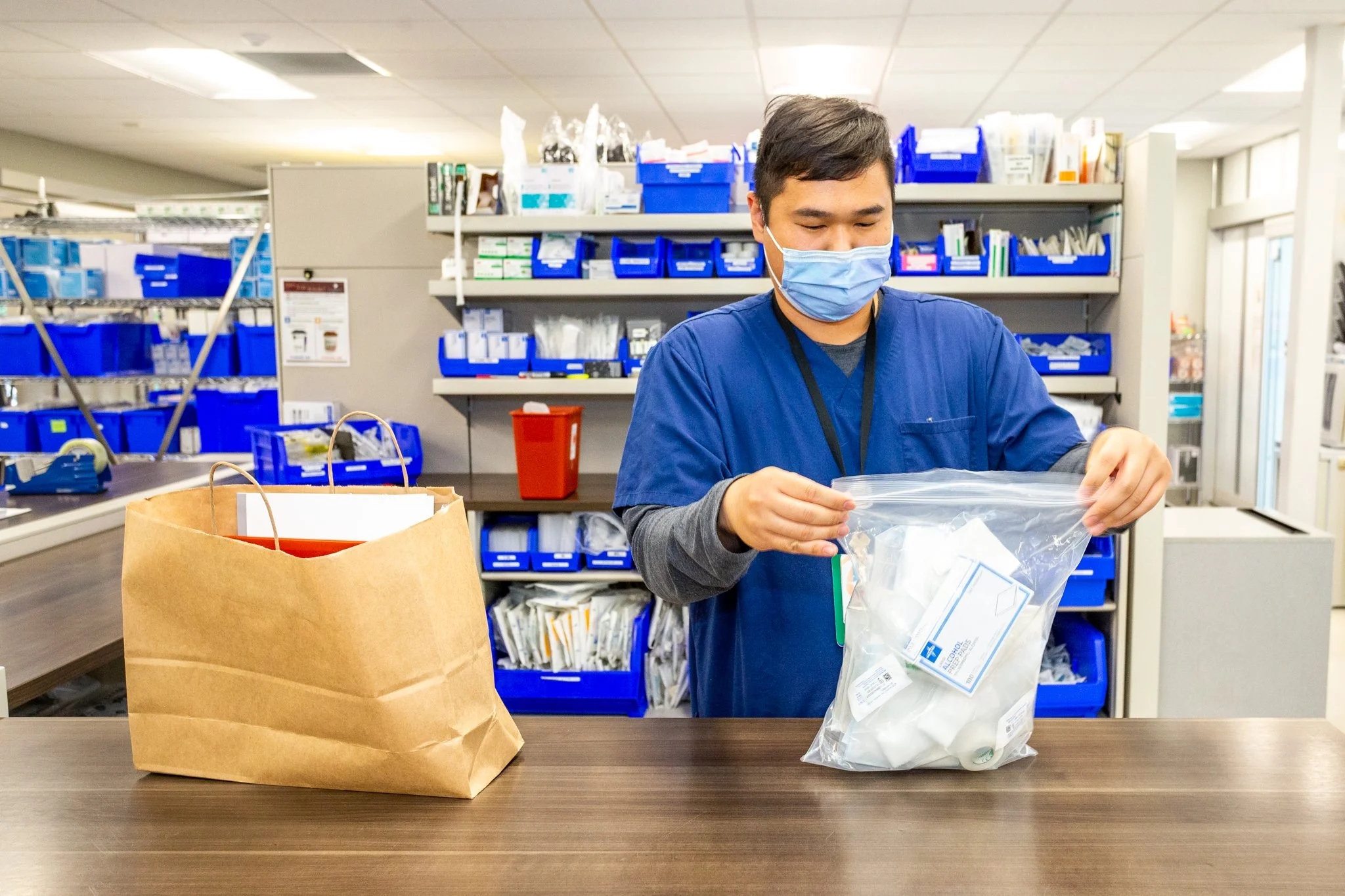 A healthcare worker wearing a face mask and blue scrubs packing medical supplies into a clear plastic bag at a hospital or clinic registration desk.