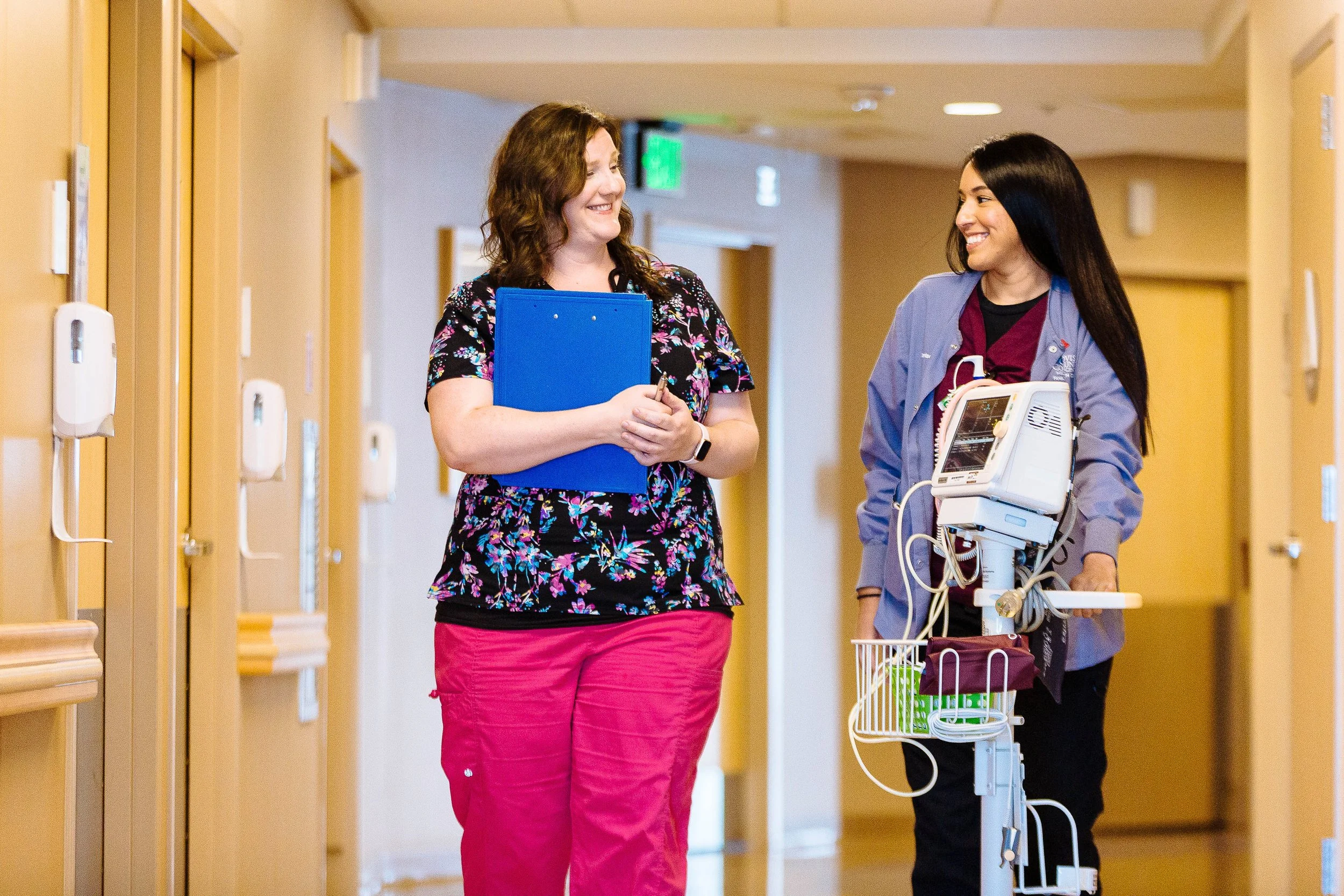A healthcare professional, wearing scrubs and standing next to a patient monitor on a rolling stand, walks down a hospital hallway talking with a visitor holding a clipboard. The hallway has beige walls and closed doors.