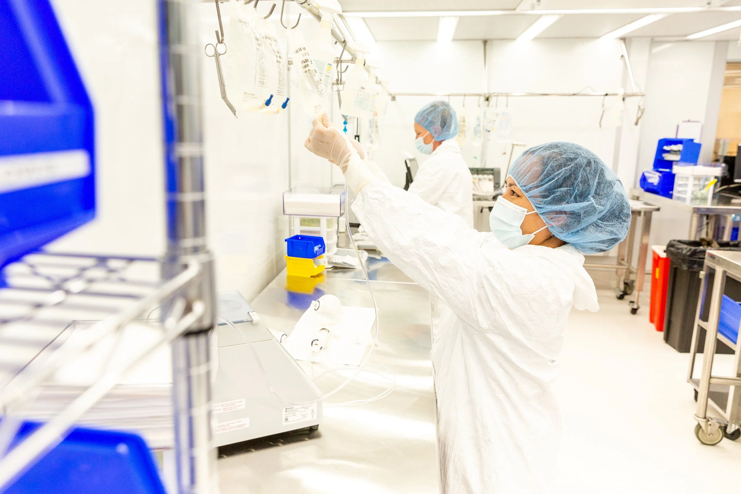 Two healthcare workers in protective gear, including surgical masks and hair covers, working in a laboratory or cleanroom environment, handling medical supplies or equipment.