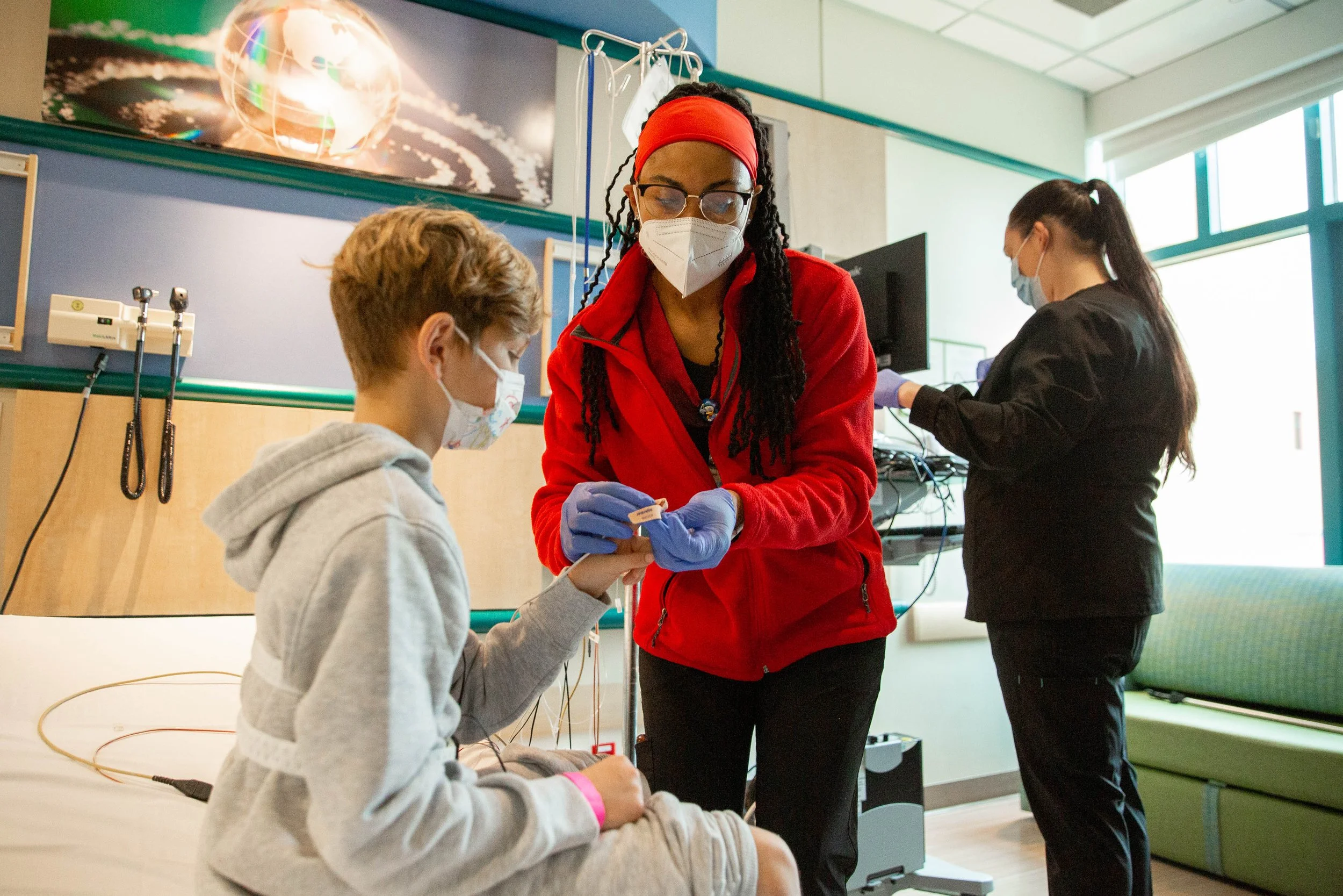 A healthcare professional in a red jacket and gloves preparing a medical injection for a young patient in a hospital room, with another healthcare worker in the background.