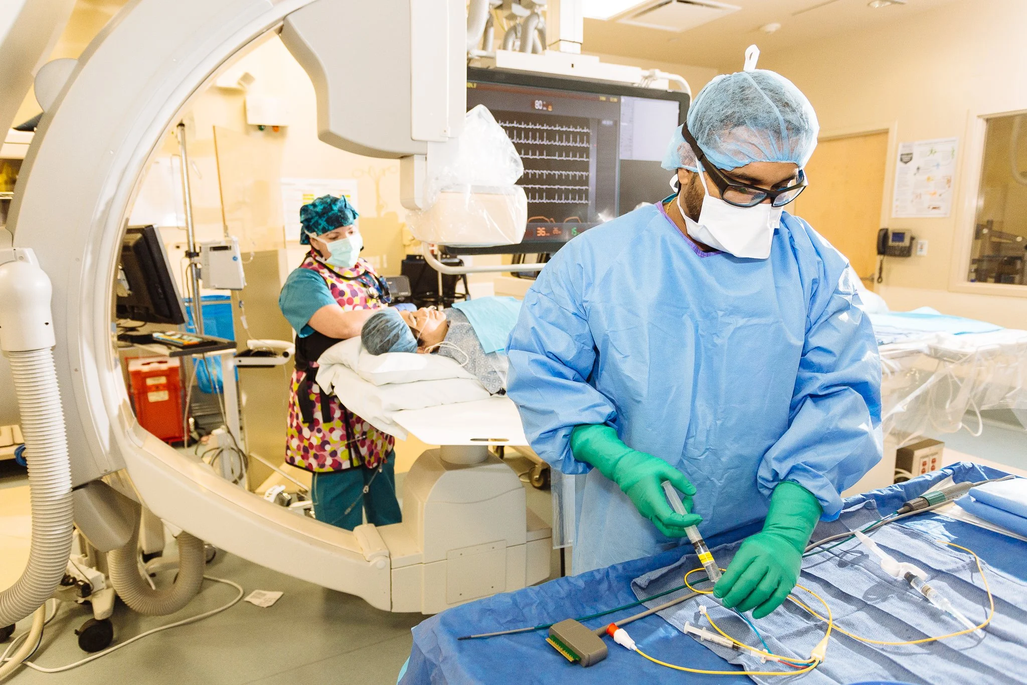 Medical professionals, including a surgeon and nurse, working in an operating room with a patient on the bed. The scene includes medical monitors, equipment, and surgical tools, with one worker preparing medical devices.