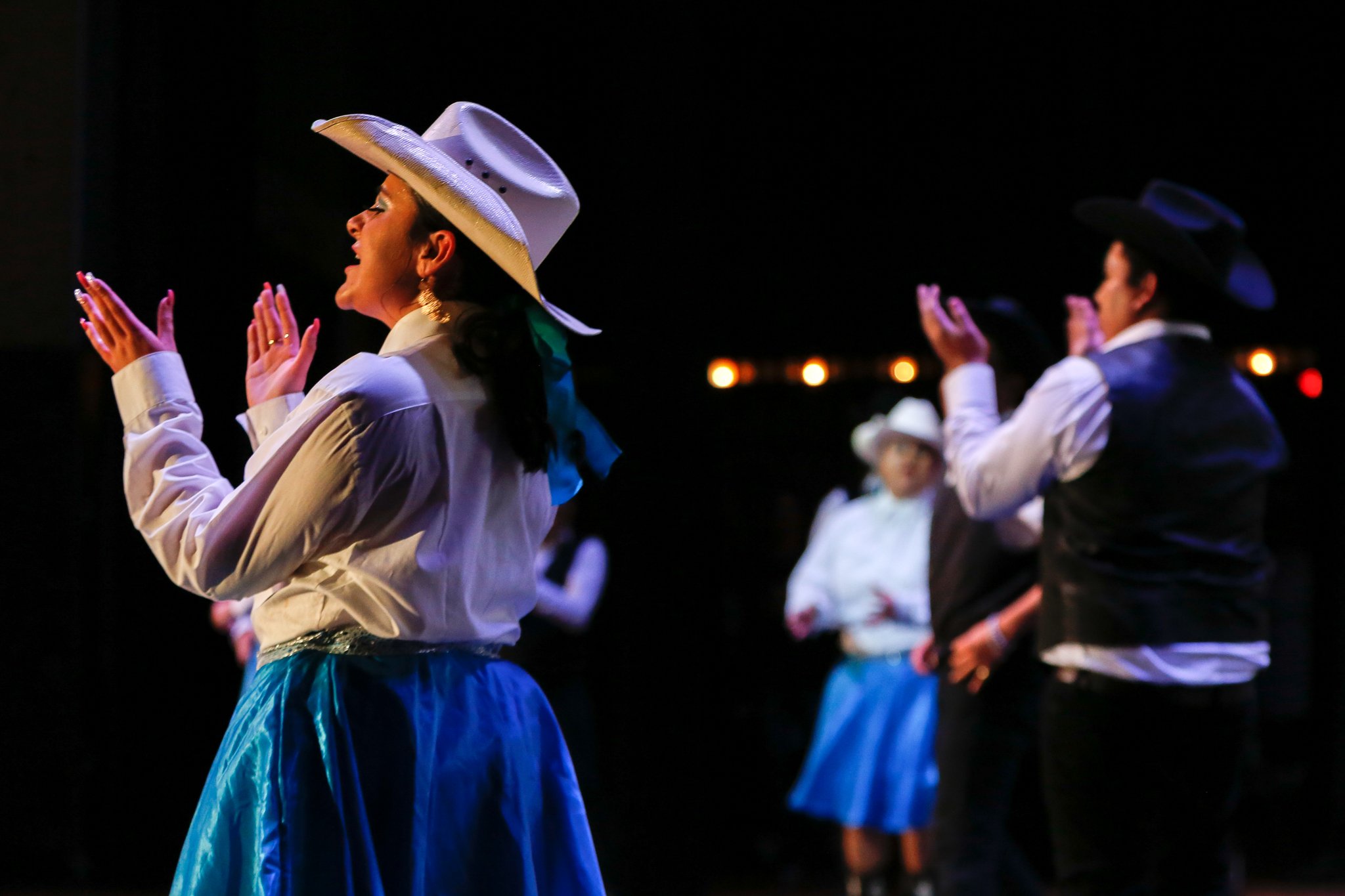 Group of people dressed in cowboy and cowgirl attire singing or performing on stage, illuminated in a dark setting.