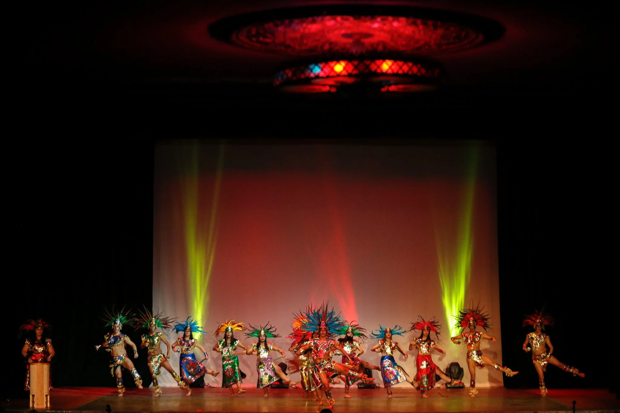 Group of dancers in vibrant costumes performing on stage with colorful feathered headpieces and energetic movements, stage lighting illuminating the background.