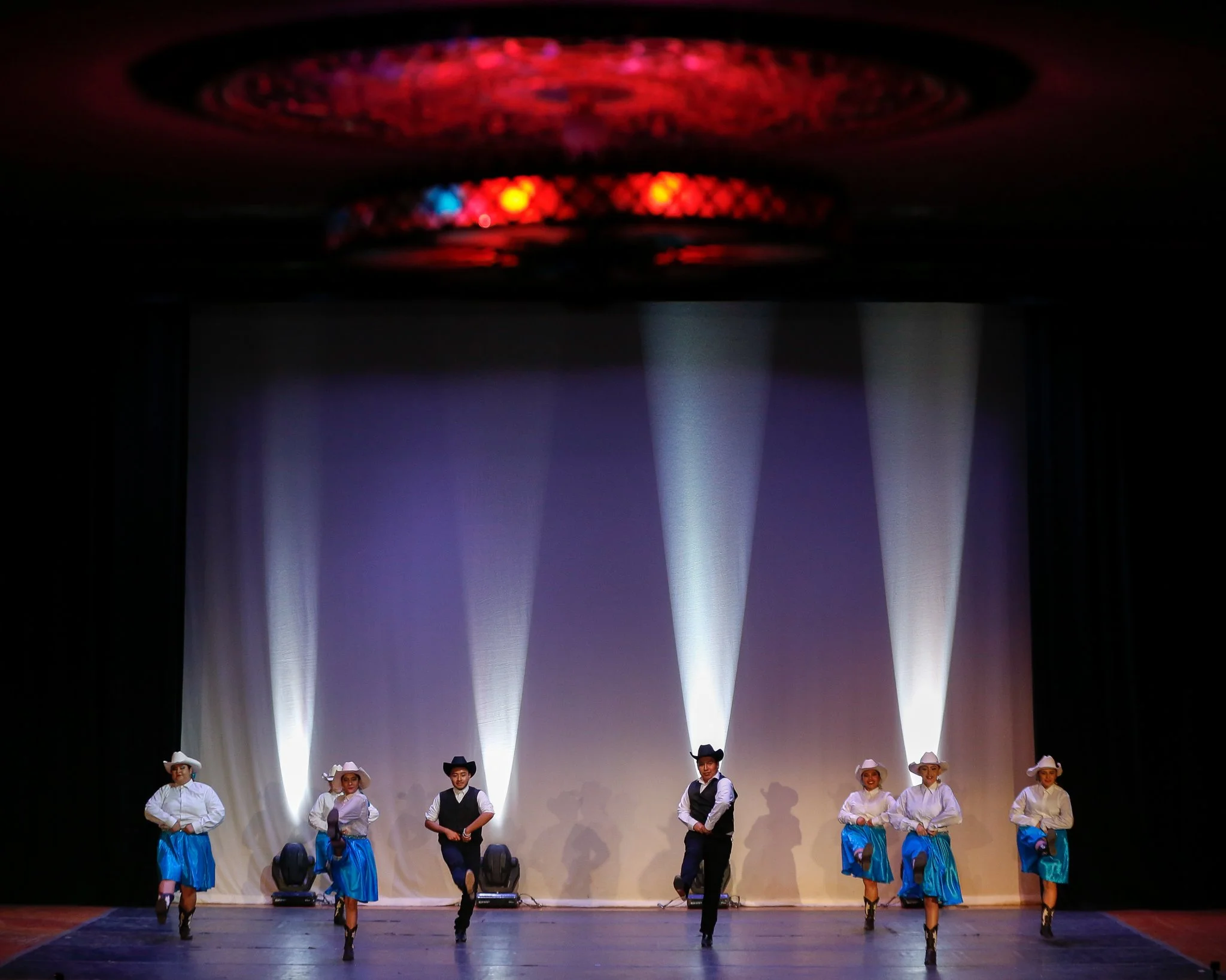 Group of six dancers in cowboy costumes performing on stage with a white curtain backdrop, colorful stage lights, and shadows, dressed in white shirts, blue skirts or vests, and cowboy hats.