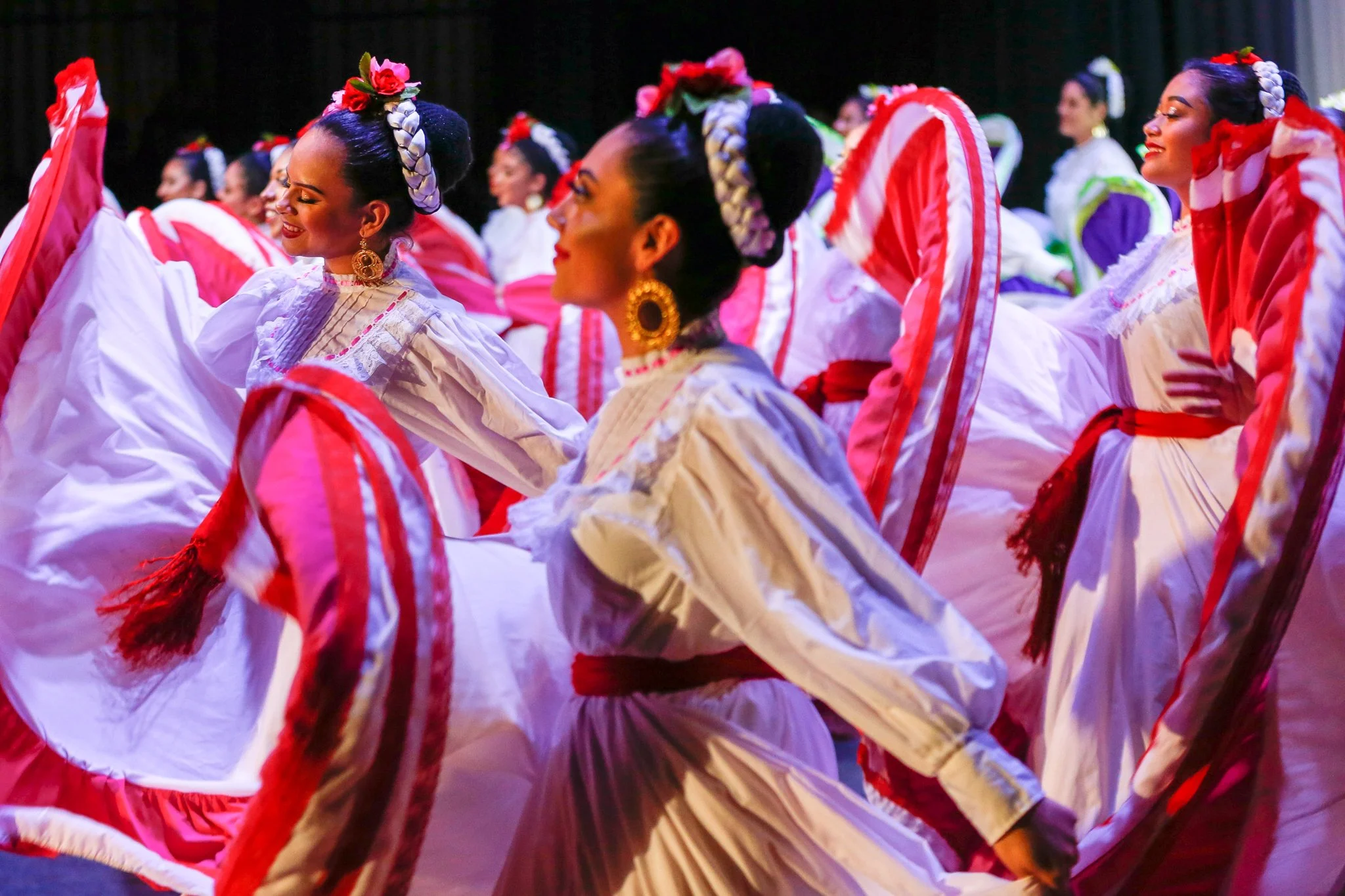 Group of women dressed in traditional Mexican folkloric dresses performing a dance, holding large colorful skirts, and wearing vibrant jewelry and flower headpieces.