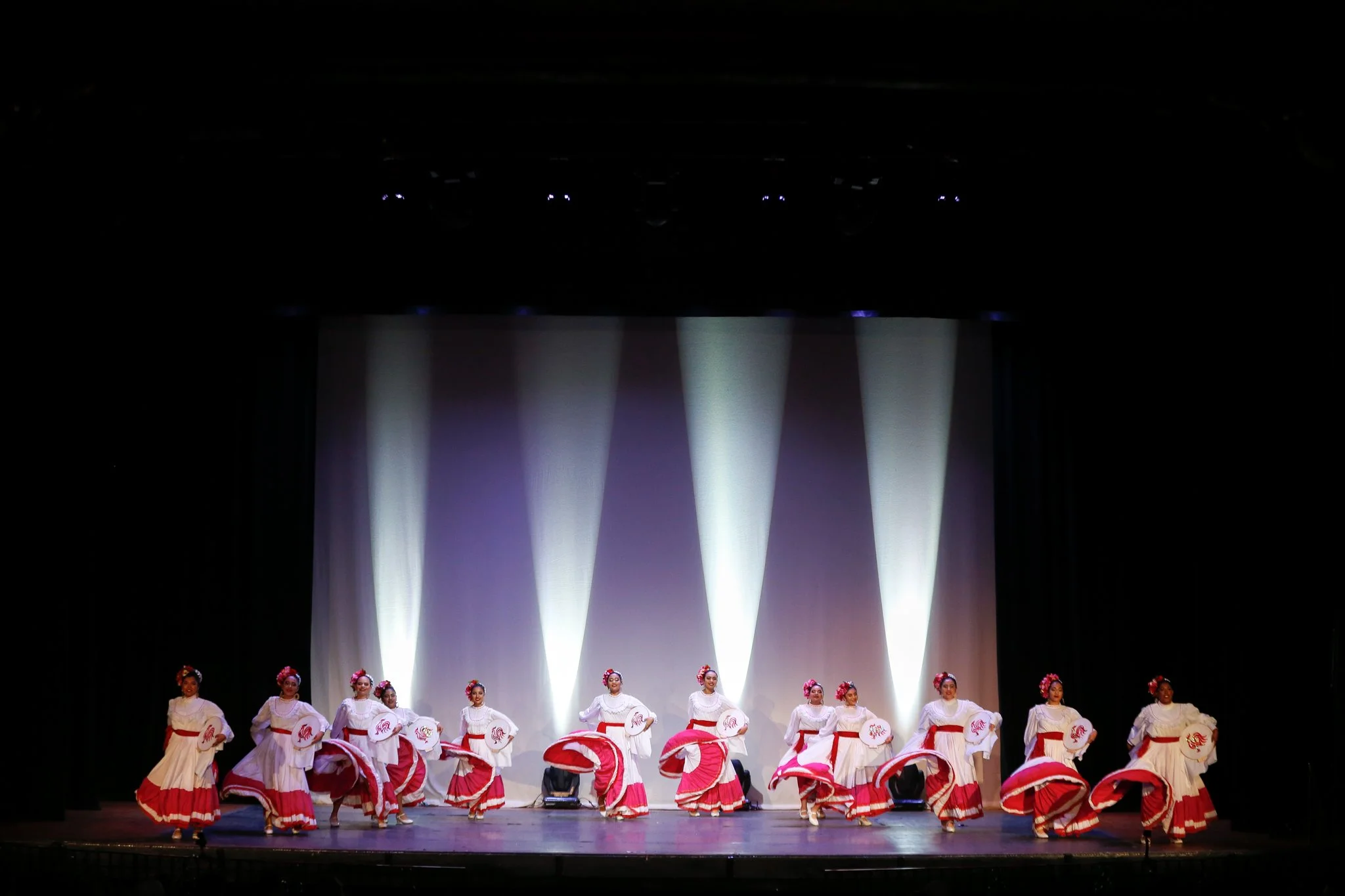 A group of ten women dressed in white and red traditional dresses performing a dance on stage, with purple and white stage lighting in the background.