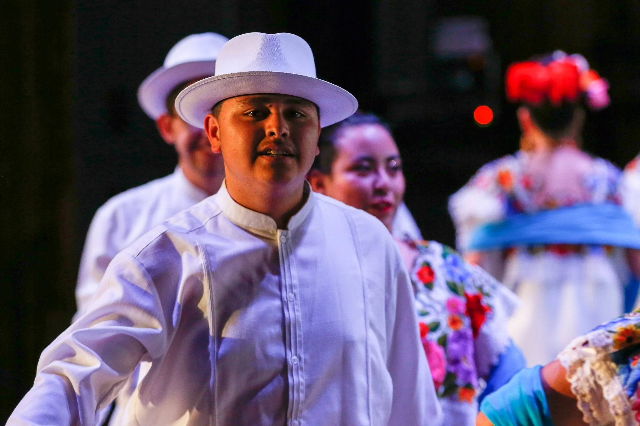 People in traditional Mexican dress, with the focus on a young man in a white shirt and wide-brimmed white hat, participating in a cultural dance or performance.