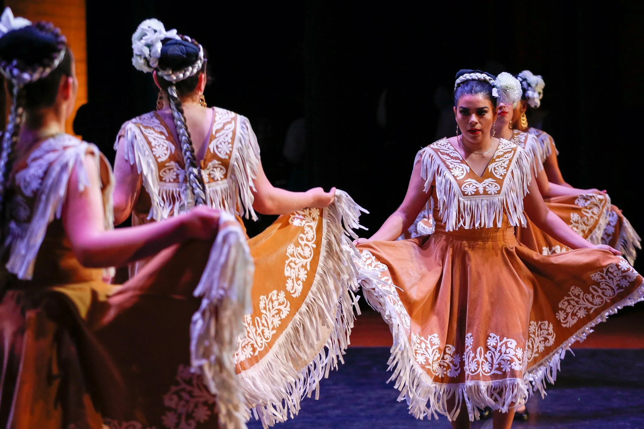 Group of women performing a traditional dance, dressed in ornate, fringed, brown and white dresses with floral embroidery, on stage with a dark background.