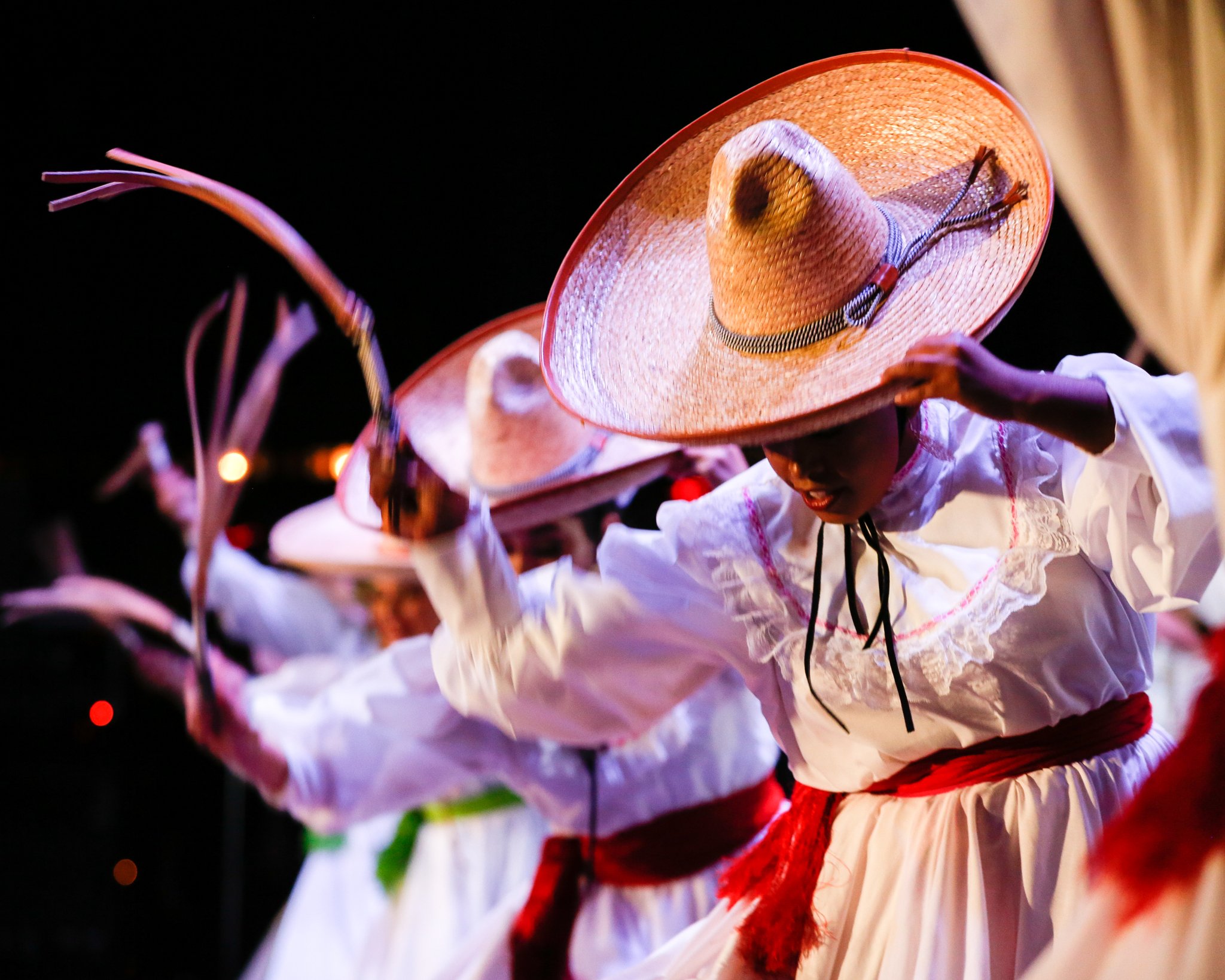 Dance performance with women dressed in traditional Mexican attire, wearing large sombreros and long white dresses with red sashes, under dark stage lighting.