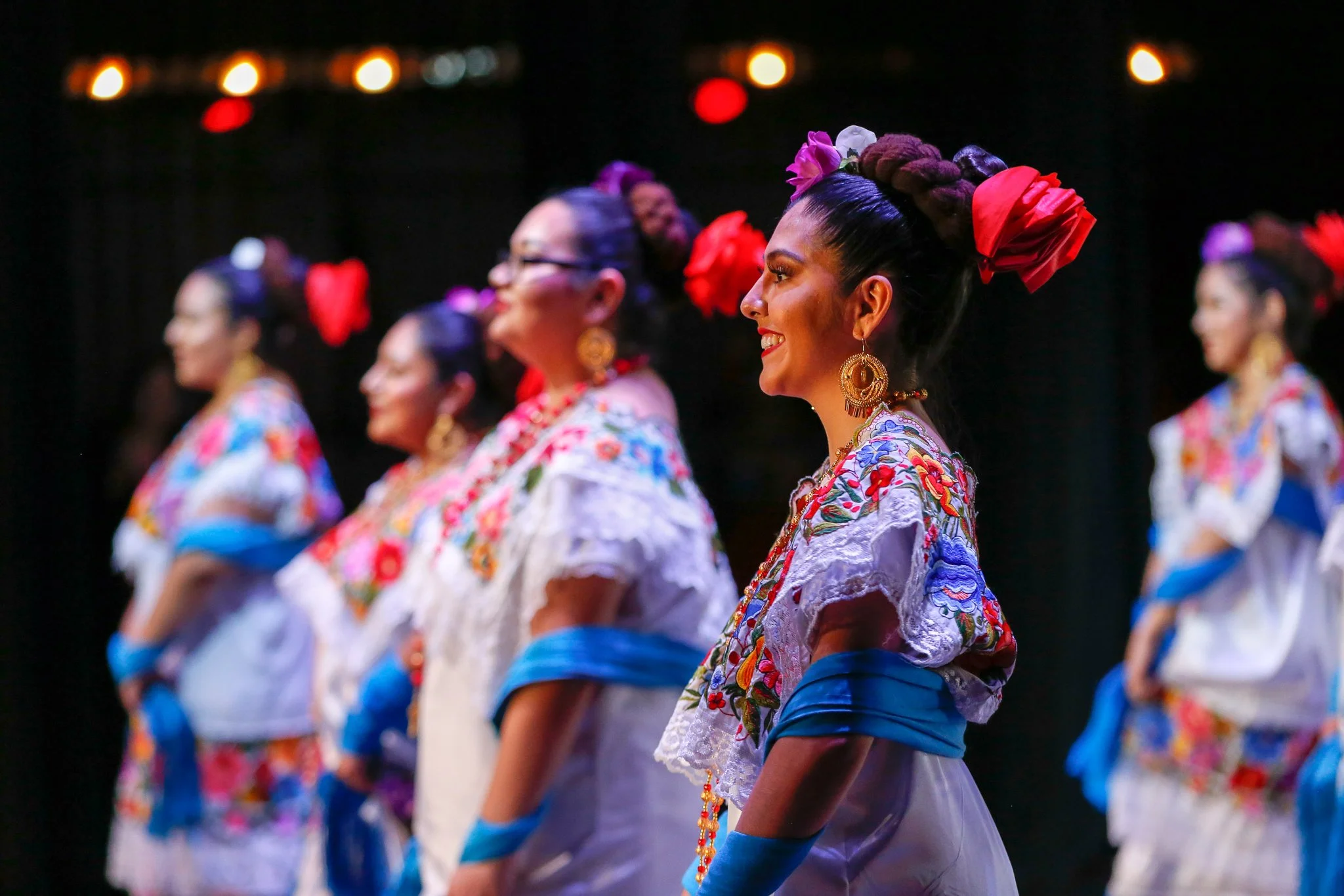Women in colorful traditional Mexican dresses dancing on stage, wearing floral embroidery, earrings, and with their hair styled in braided updos adorned with large red and purple flowers.