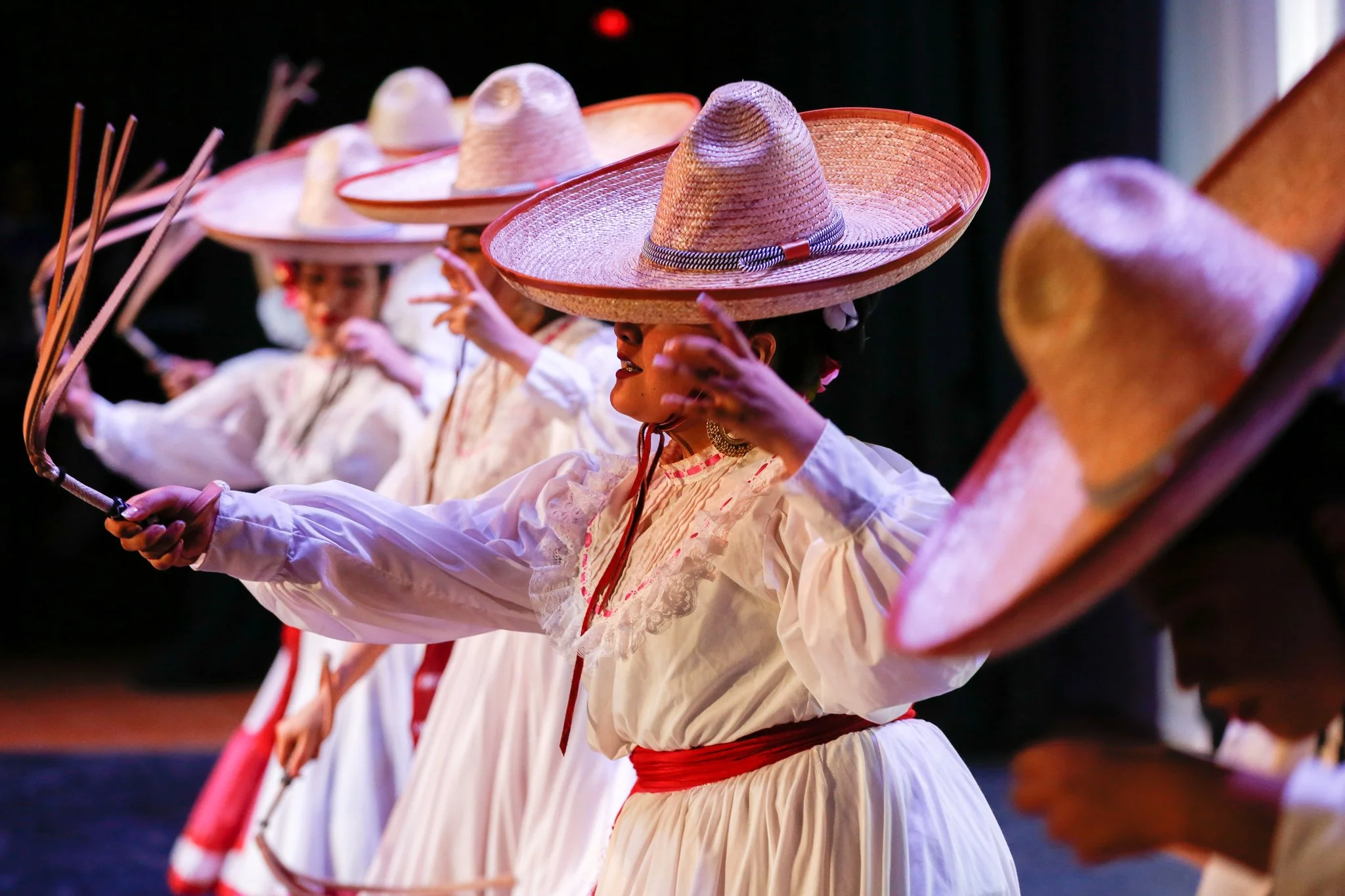 Mexican folkloric dancers wearing traditional white dresses and large sombrero hats, performing with wooden sticks.