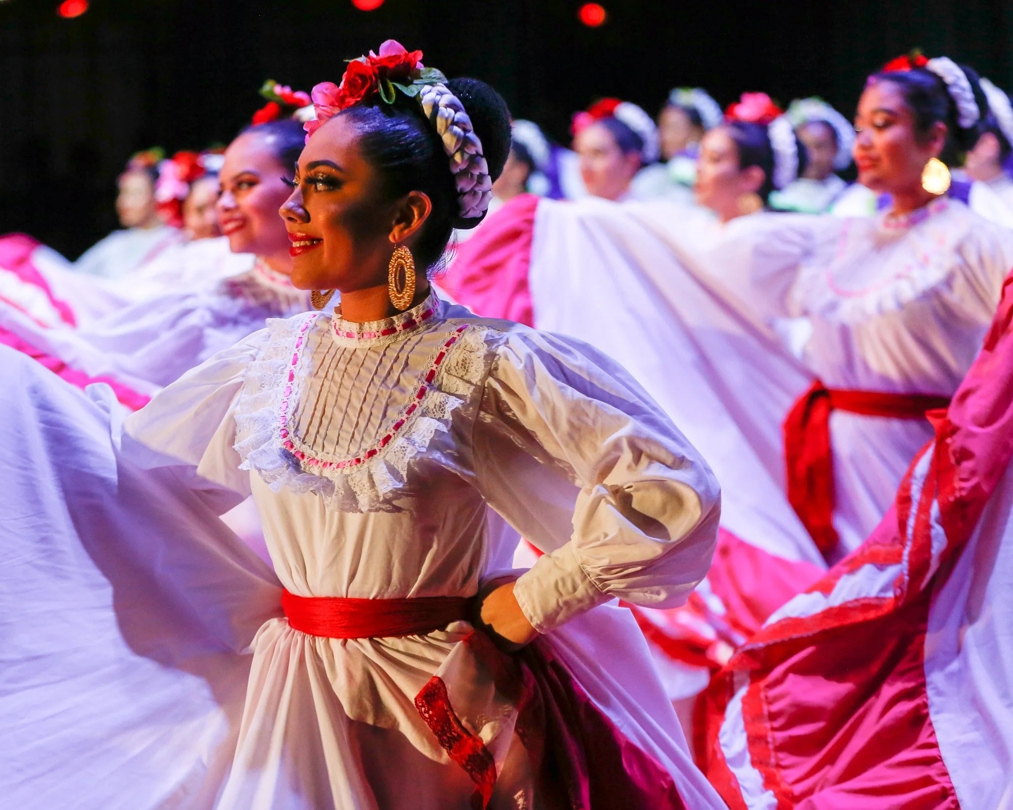 Group of women dressed in traditional colorful Mexican folkloric dresses, performing a dance, with the focus on a smiling woman in the foreground.
