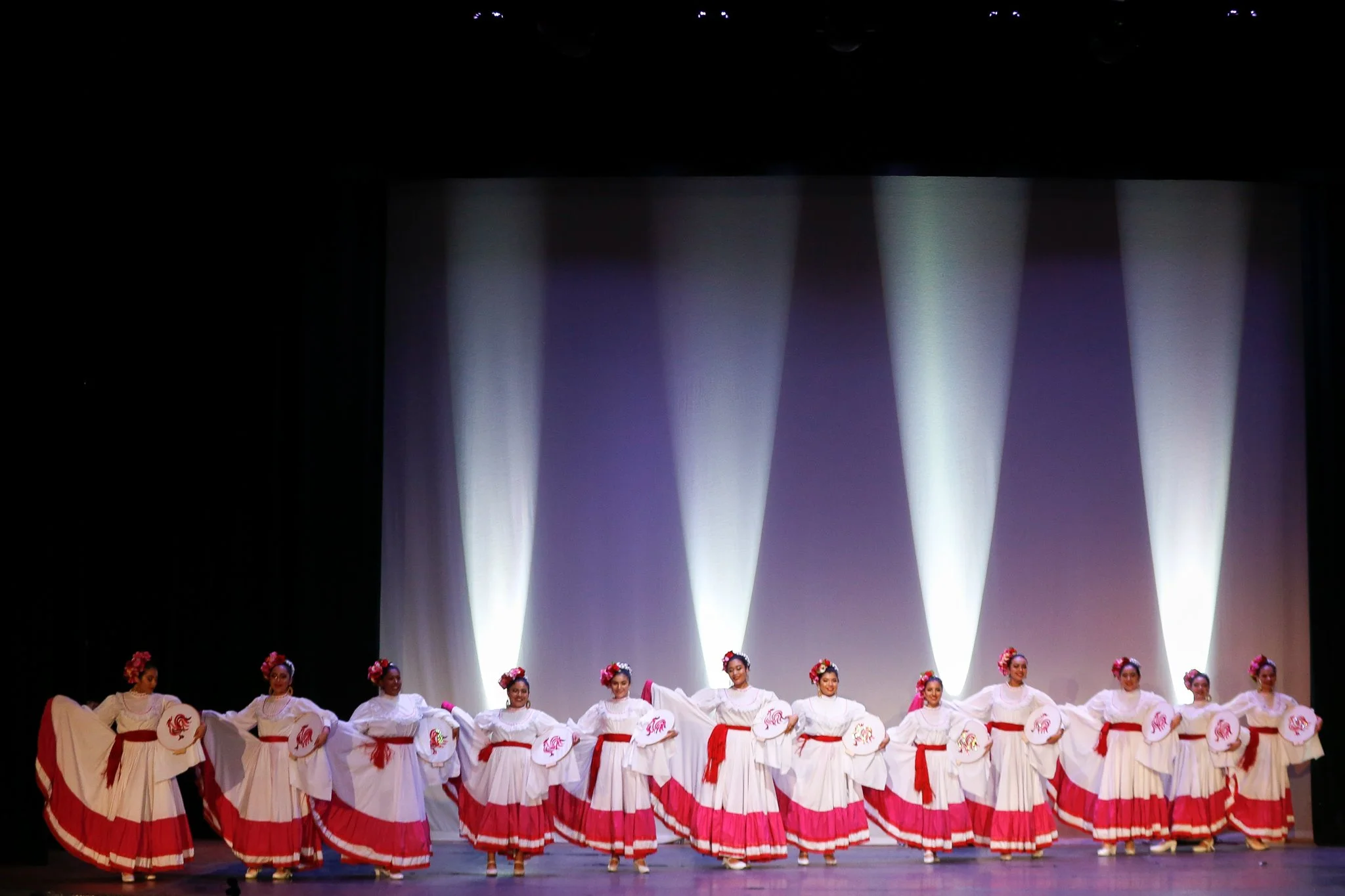 A group of dancers dressed in white and red costumes performing a traditional dance on stage, with spotlight effects in the background.