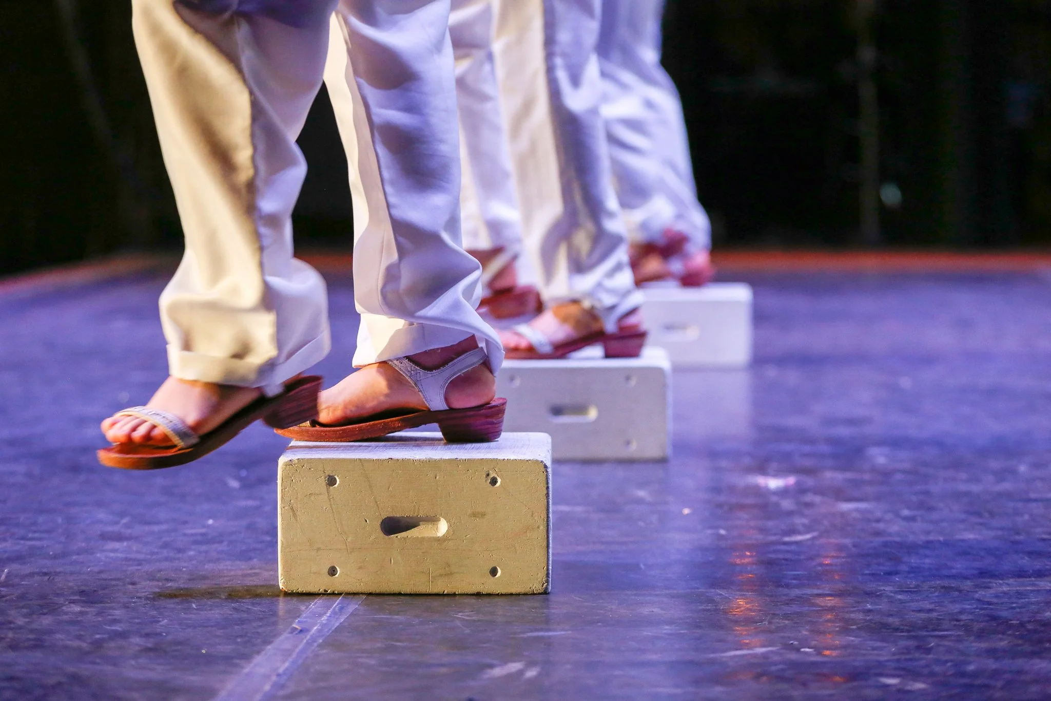 A line of dancers' legs and feet balancing on wooden blocks on stage, wearing traditional Japanese footwear and white garments.