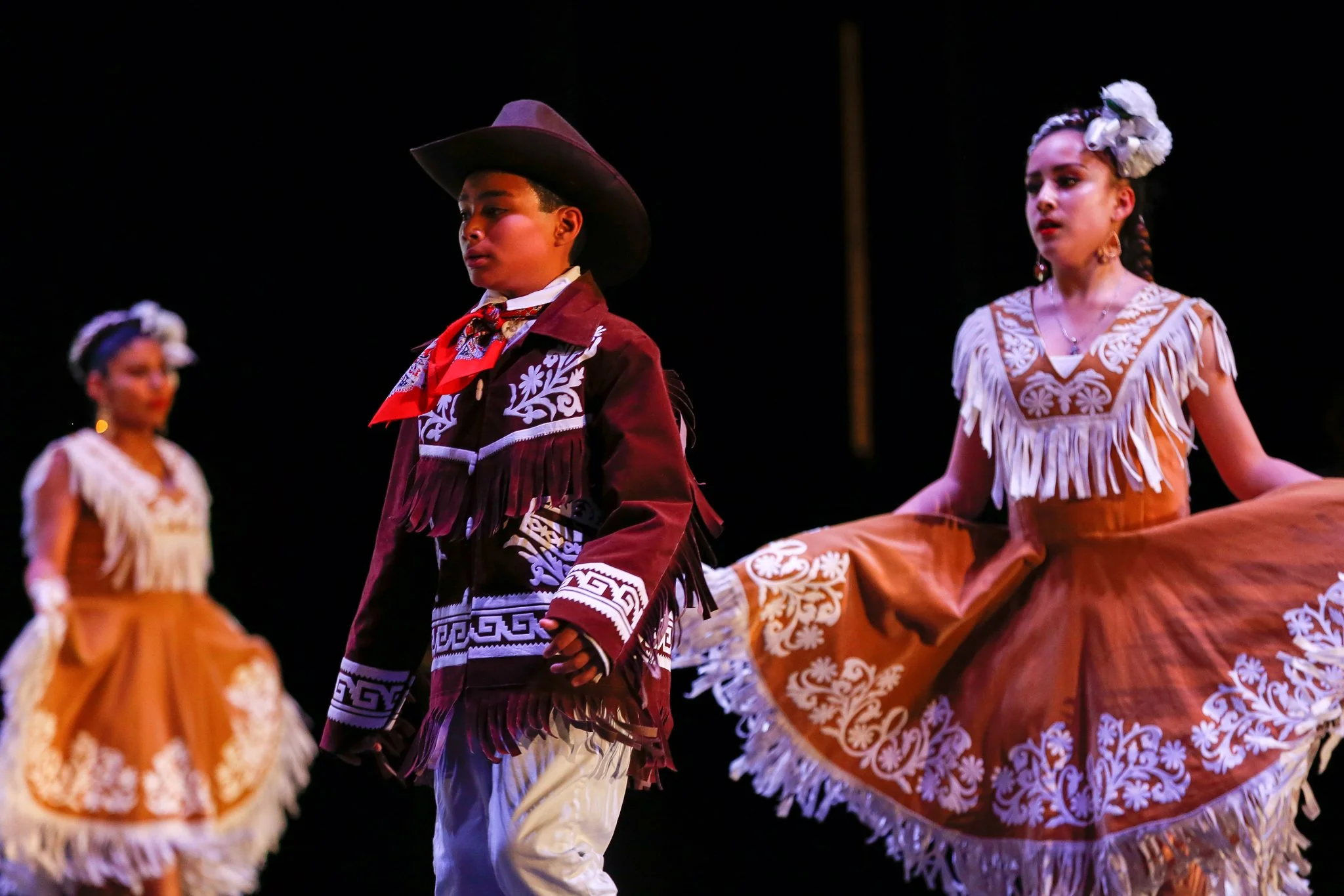 Children dressed in traditional Mexican folkloric costumes performing a dance on stage.