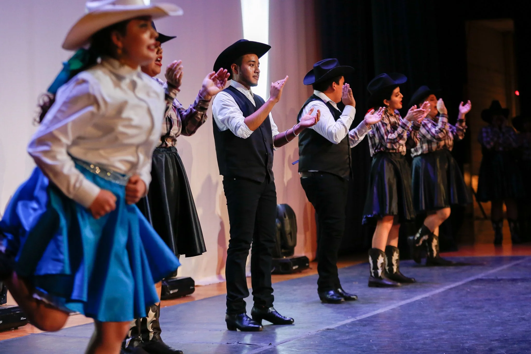 Group of dancers in cowboy costumes performing on stage, wearing cowboy hats, plaid shirts, western skirts, and boots.