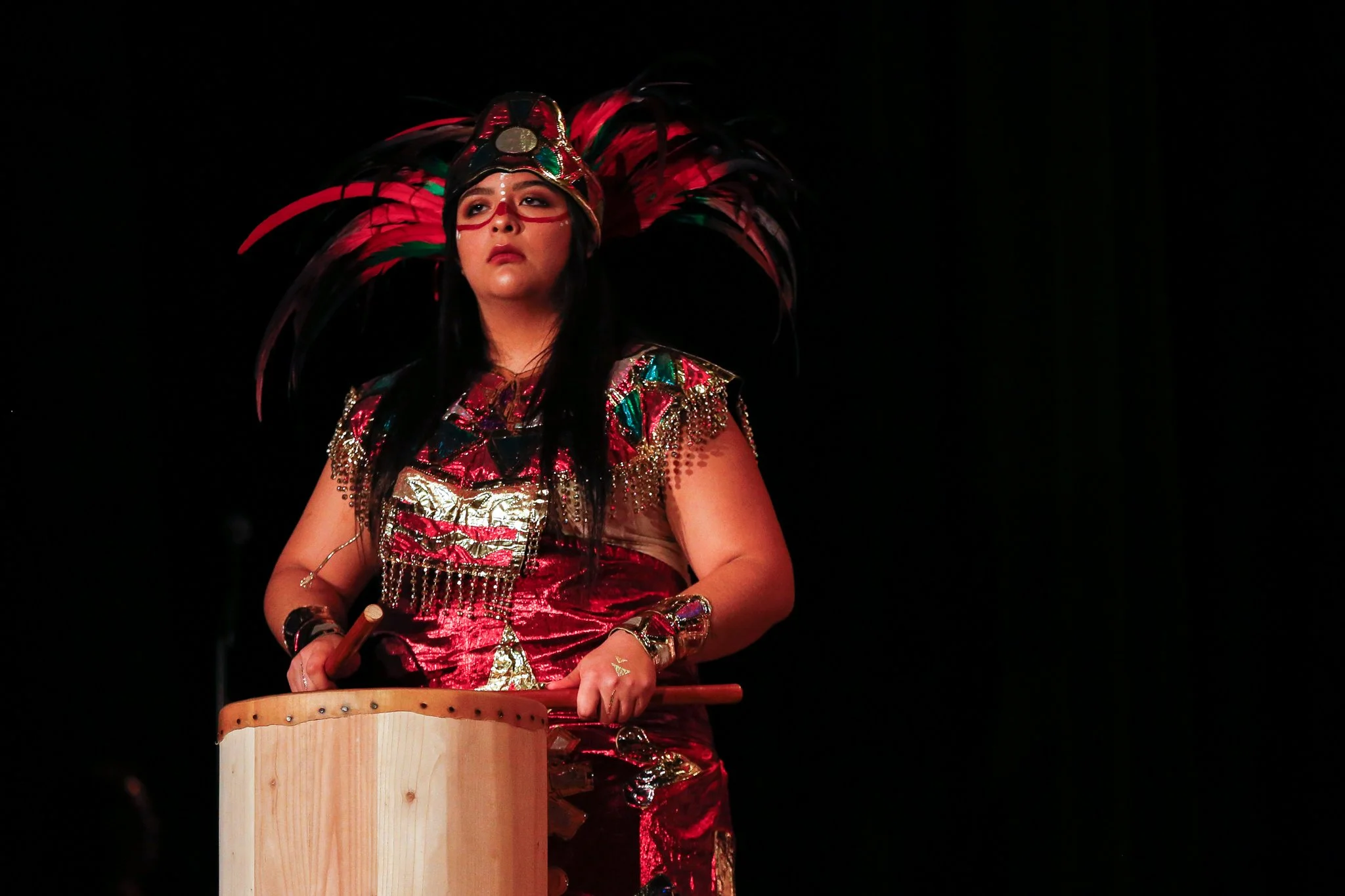 Woman dressed in an elaborate, colorful costume with feathers, metallic embellishments, and face paint, playing a large drum on stage
