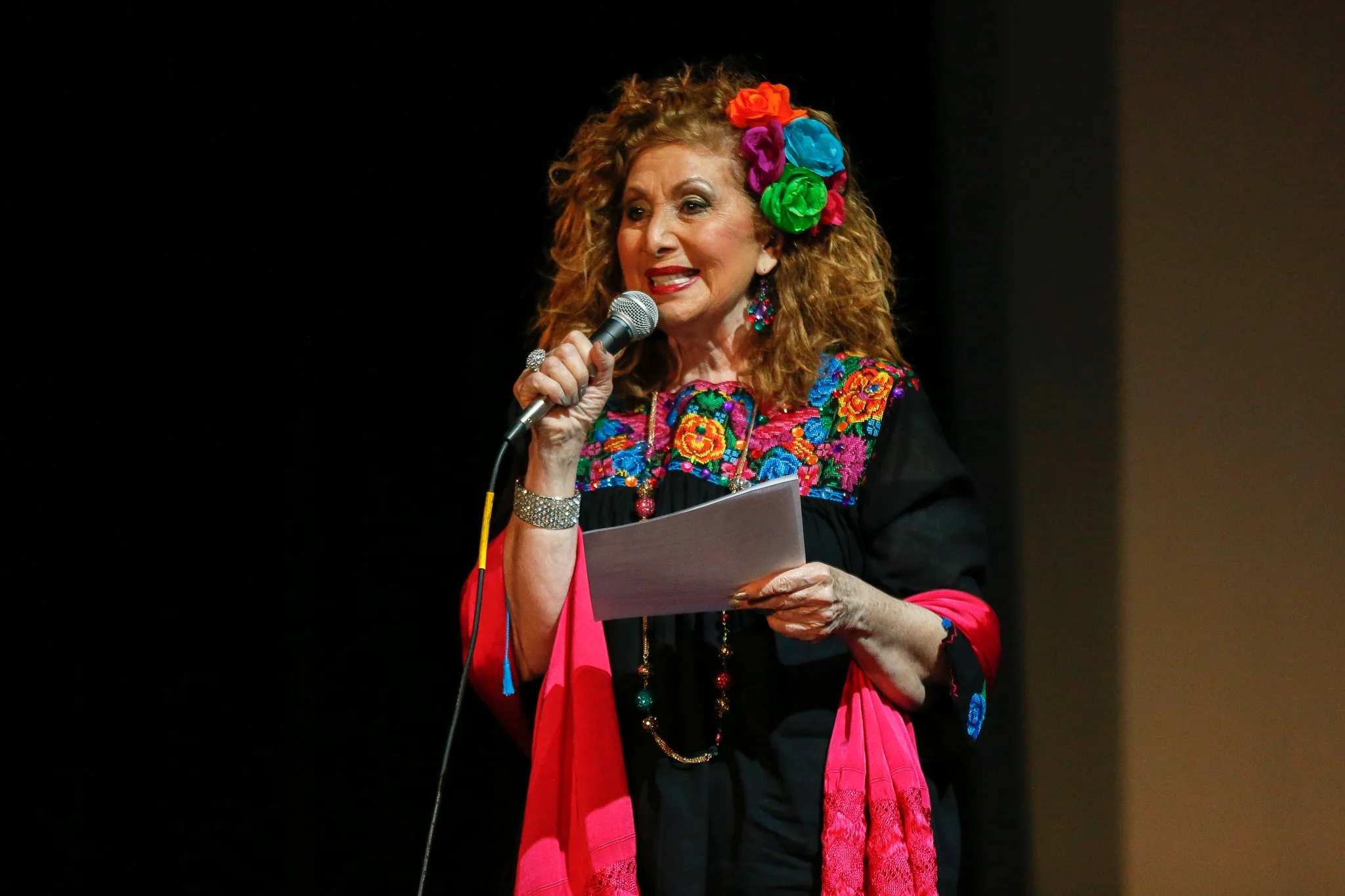 An older woman with curly red hair, wearing colorful flowers in her hair and a multicolored embroidered blouse, speaking into a microphone on a dark stage.