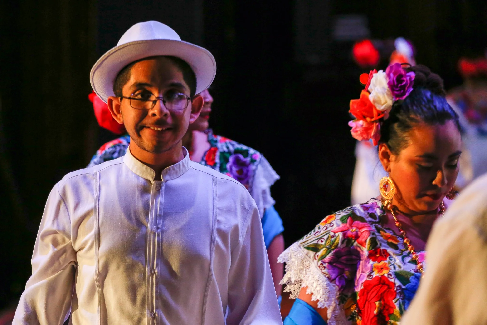 People dressed in traditional Mexican clothing, including a man in a white charro suit and hat, and women with colorful floral dresses and accessories, possibly participating in a folkloric dance or celebration.