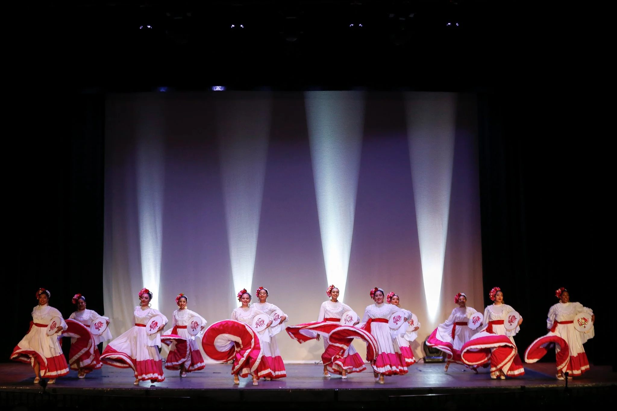 Group of women in white and red traditional dresses performing a dance on stage with spotlight effects.