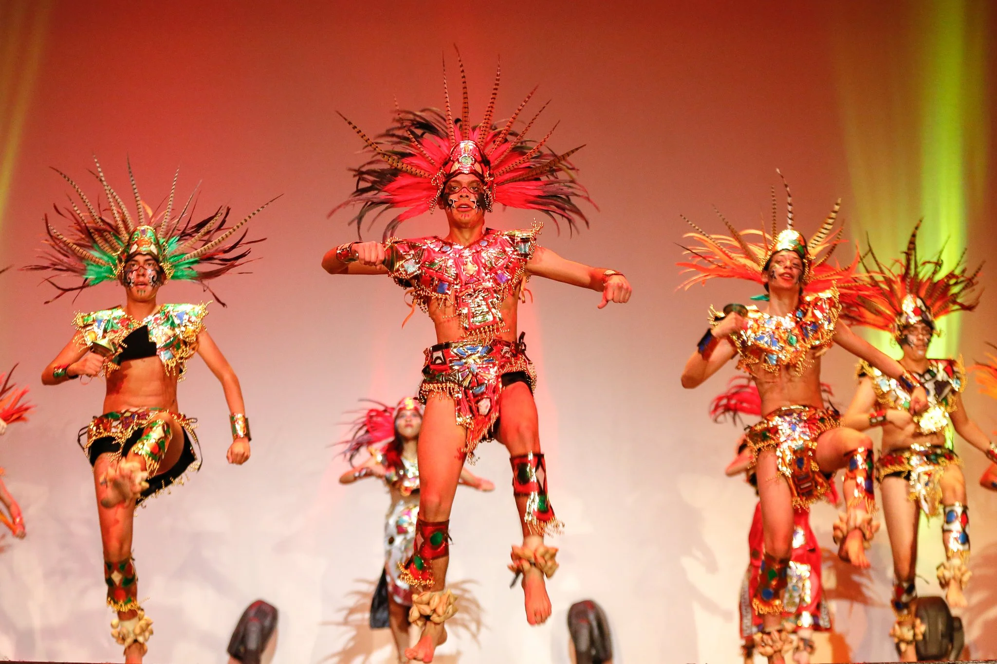 A group of dancers performing a traditional dance on stage, wearing colorful costumes with elaborate feathered headpieces.