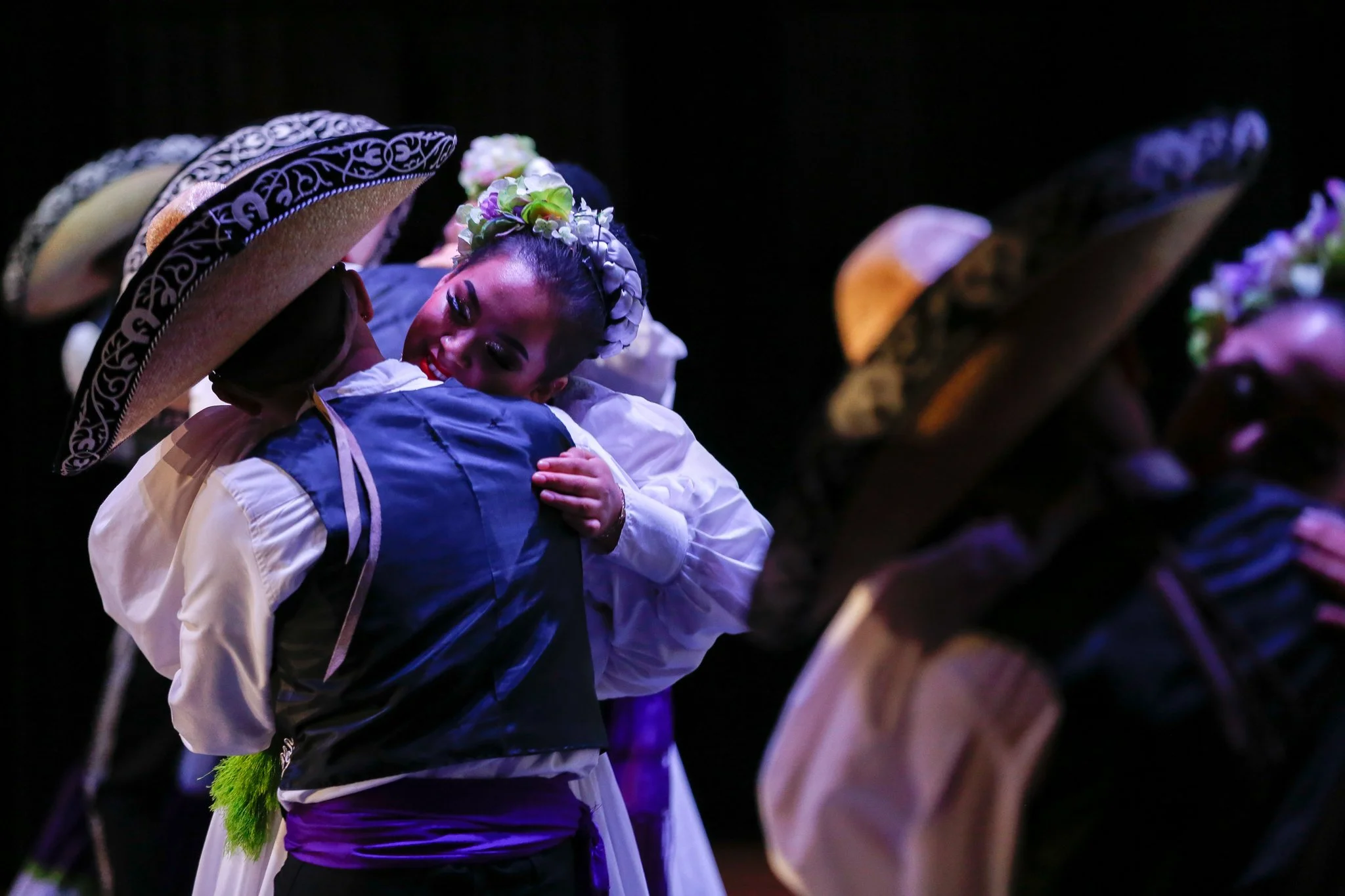 A woman dressed in traditional Mexican attire hugging a man dressed in similar traditional clothing, both wearing large sombreros. The woman is smiling and wrapped in a flower crown, while they embrace.