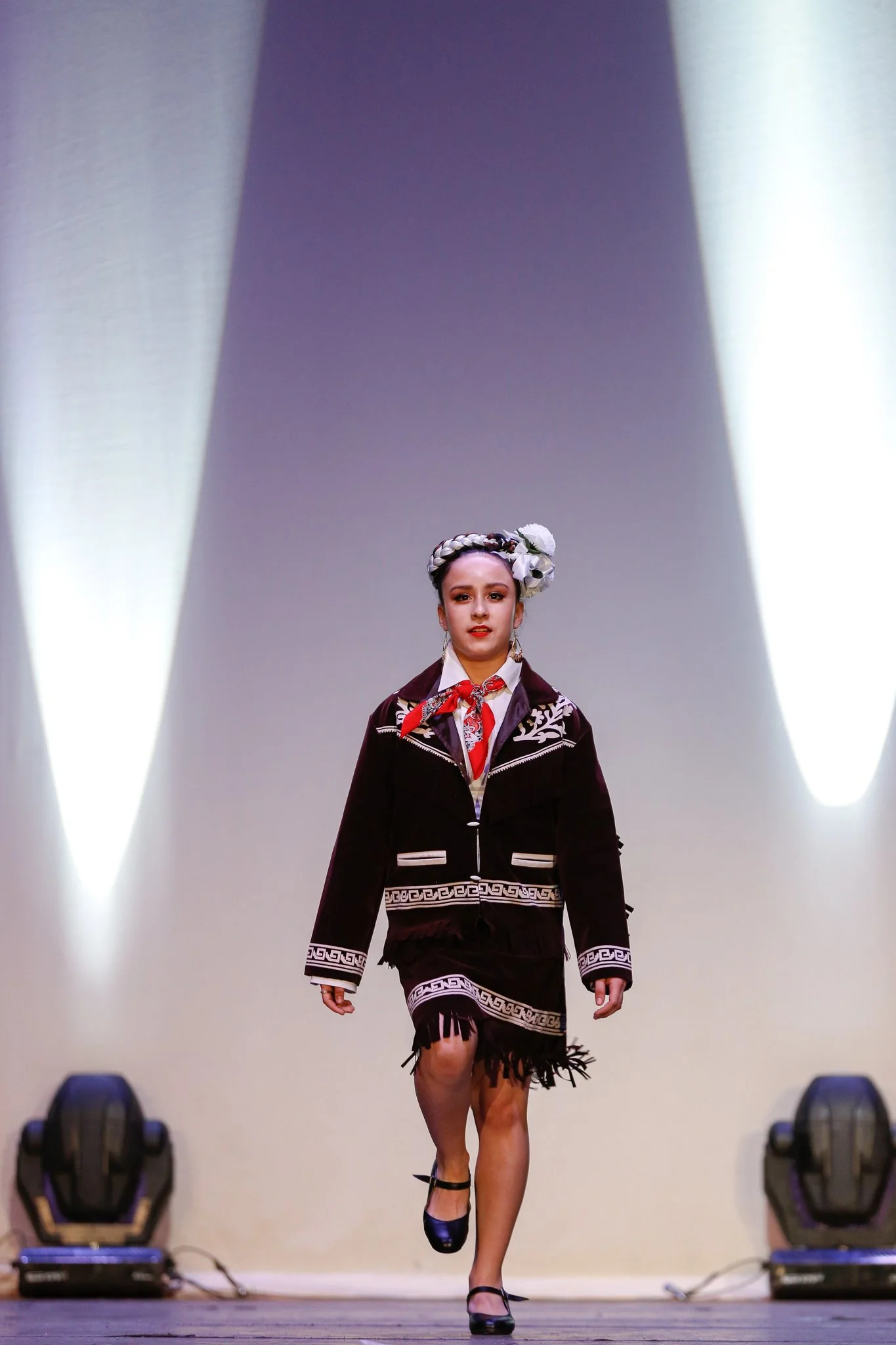 A young girl walking on a runway, wearing traditional clothing with black shoes and a floral headpiece, under stage lighting.