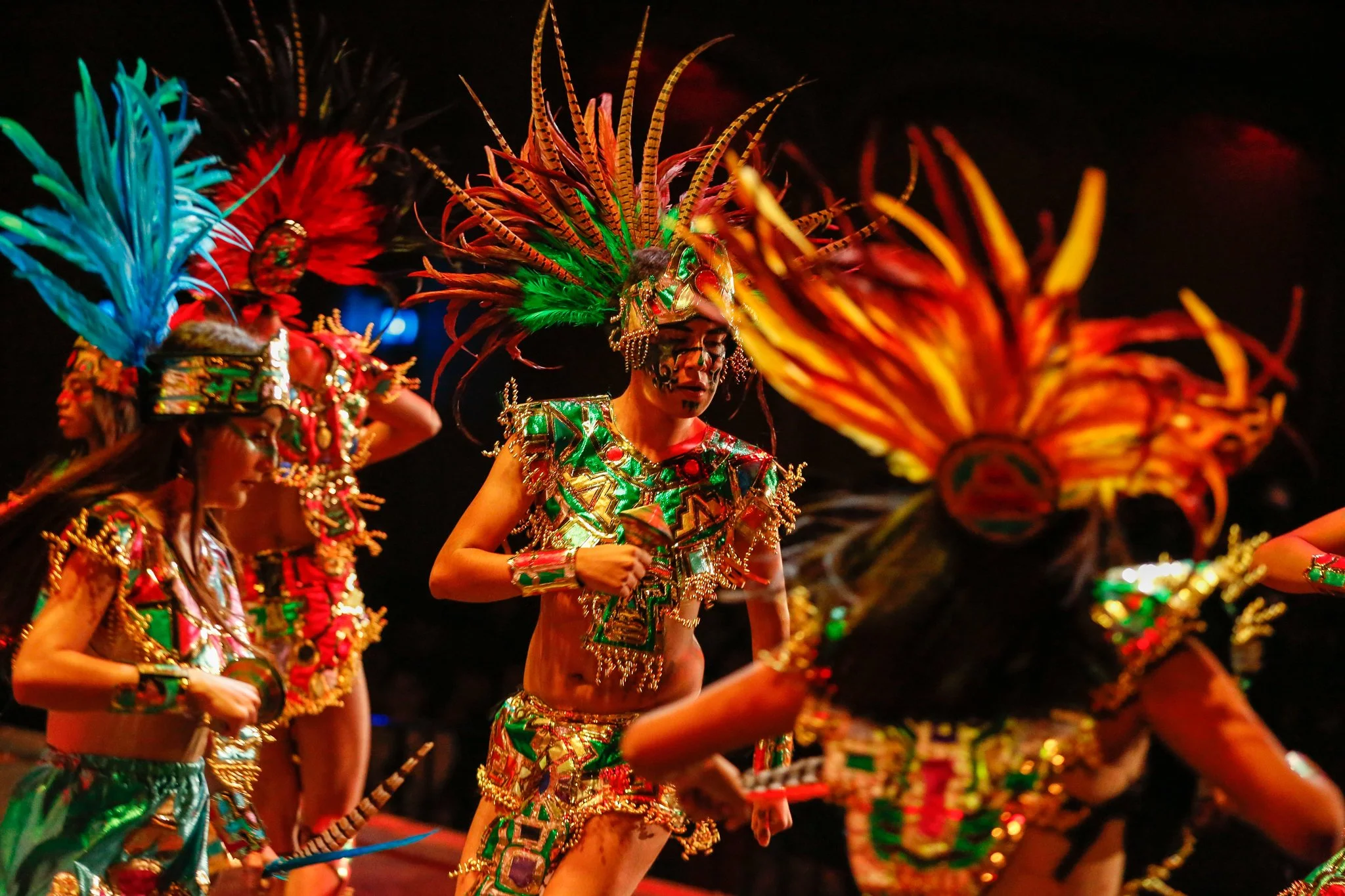 Group of performers in colorful costumes and feathered headdresses dancing on stage at night.