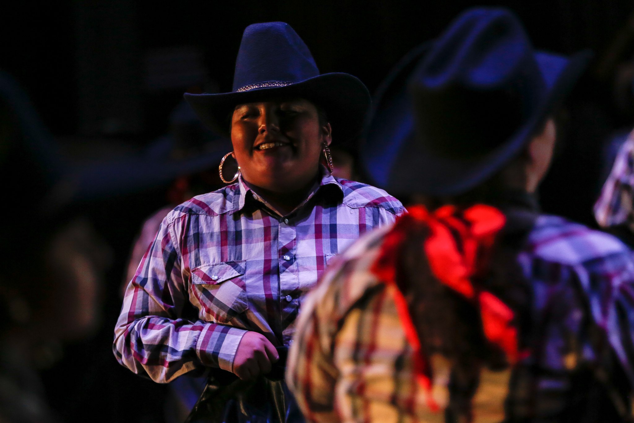 A woman with hoop earrings and a wide-brimmed hat at a nighttime event, smiling and wearing a plaid shirt, among others also wearing similar hats and plaid shirts.