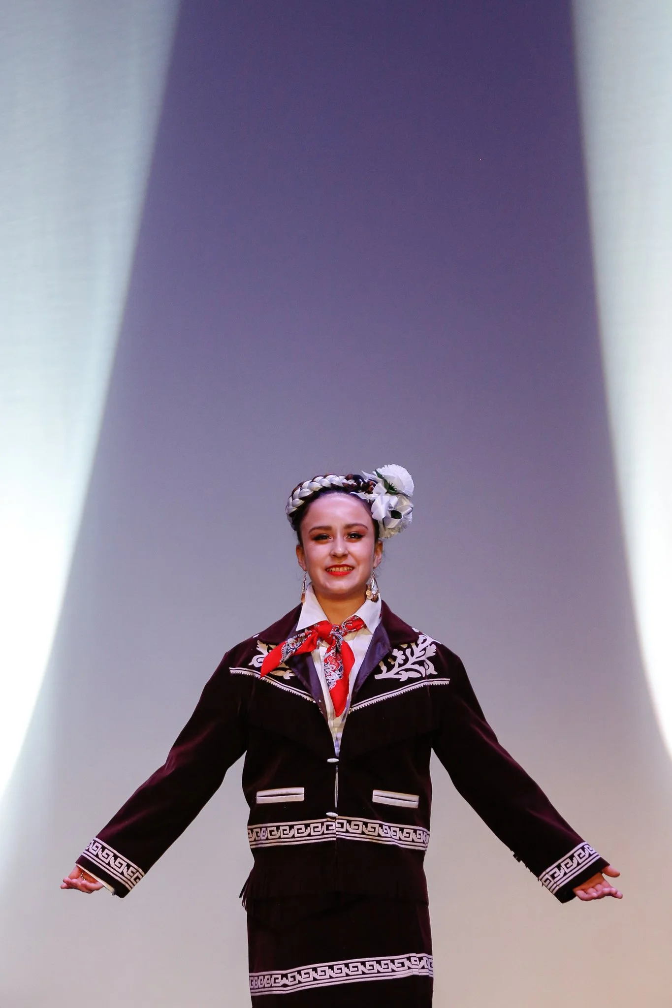 A woman dressed in traditional attire, smiling, with her arms extended to the sides, standing on a stage with a plain background.
