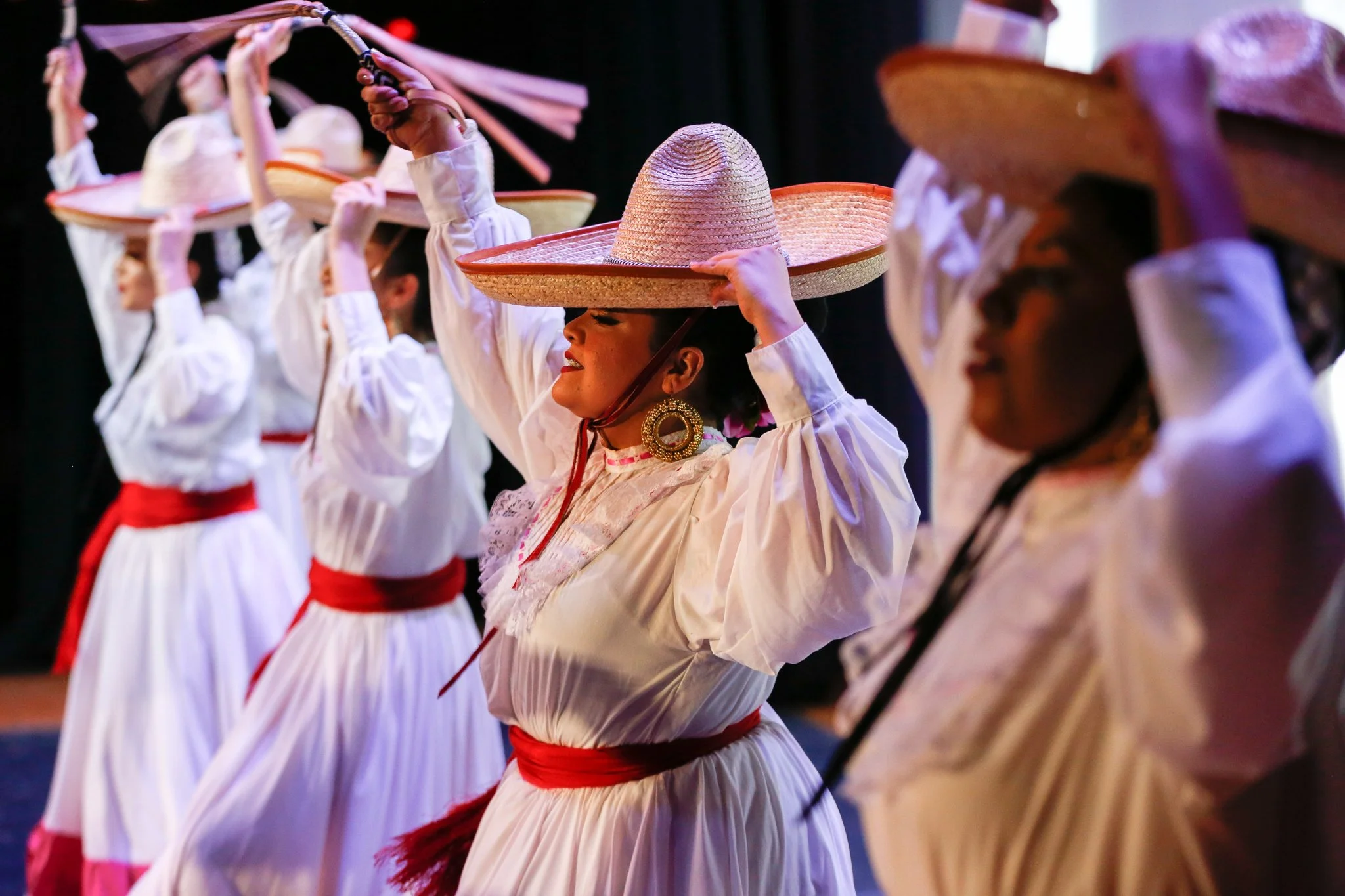 A group of women dressed in traditional white dresses with red sashes, wearing large straw sombreros, and performing a dance on stage.