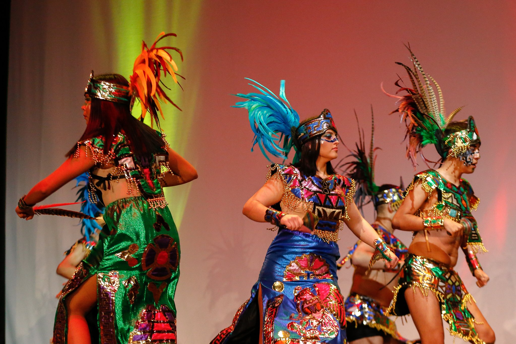 Group of women wearing colorful traditional costumes with feathers and jewelry, performing a dance on stage.