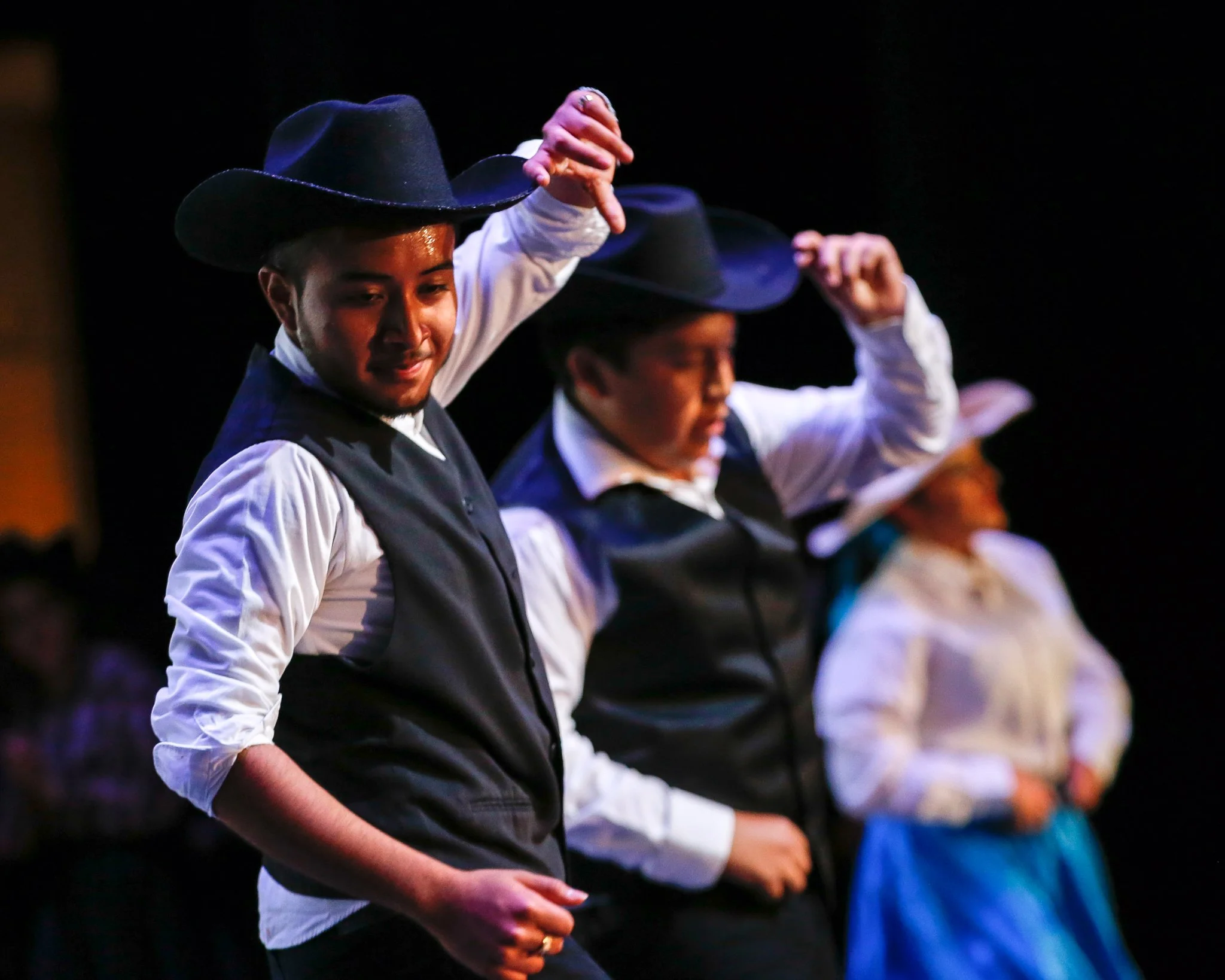 Two men dressed in cowboy attire, wearing black hats, white shirts, and vests, are dancing on stage with a woman in the background, also in vintage outfit, under stage lighting.