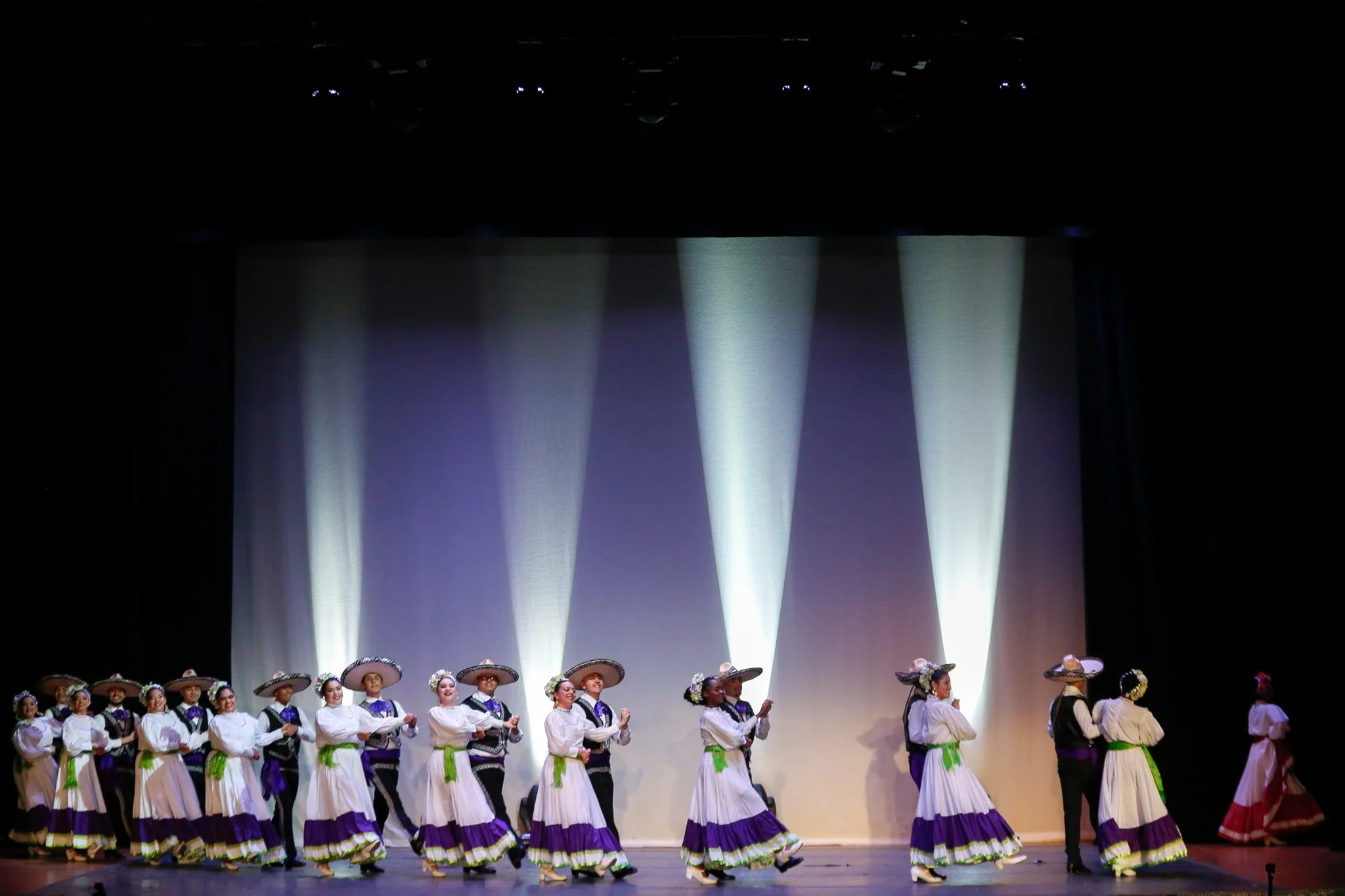A group of dancers in traditional Mexican folkloric costumes performing on a stage with white and purple dresses and large sombreros, lit by stage lights.
