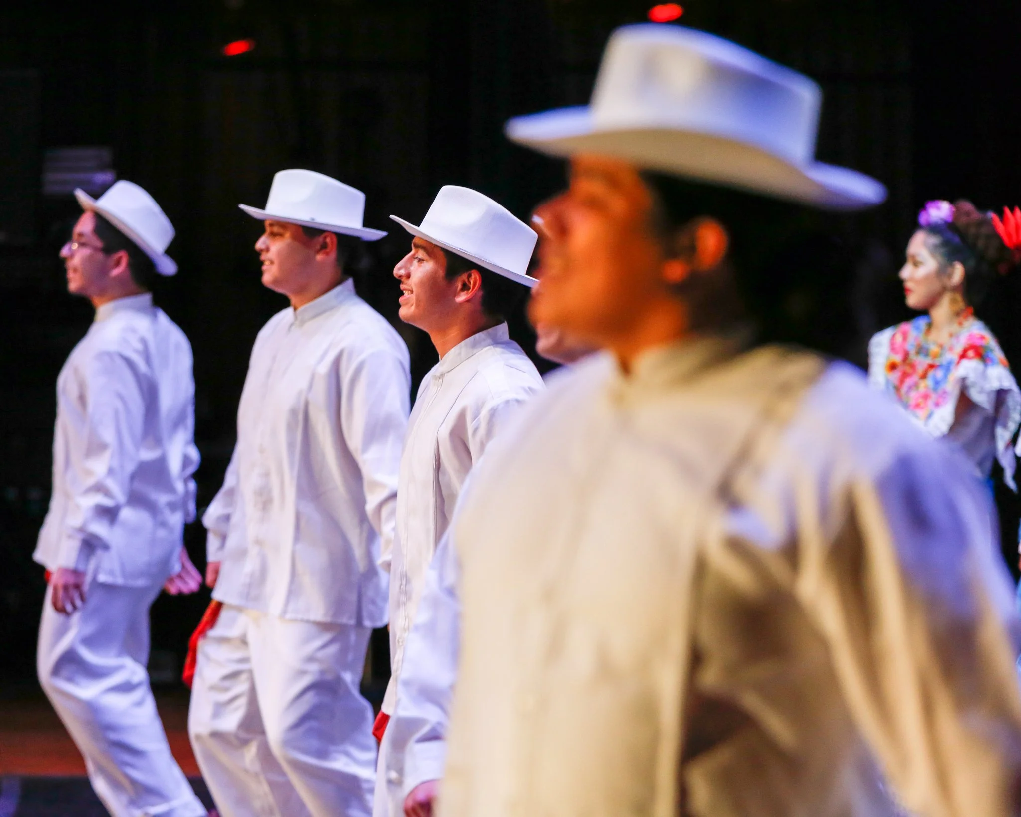 A group of five dancers dressed in traditional Mexican attire, wearing white outfits and wide-brimmed hats, performing on stage with a dark background. The woman in the background is wearing a colorful dress with floral patterns and a pink flower in 