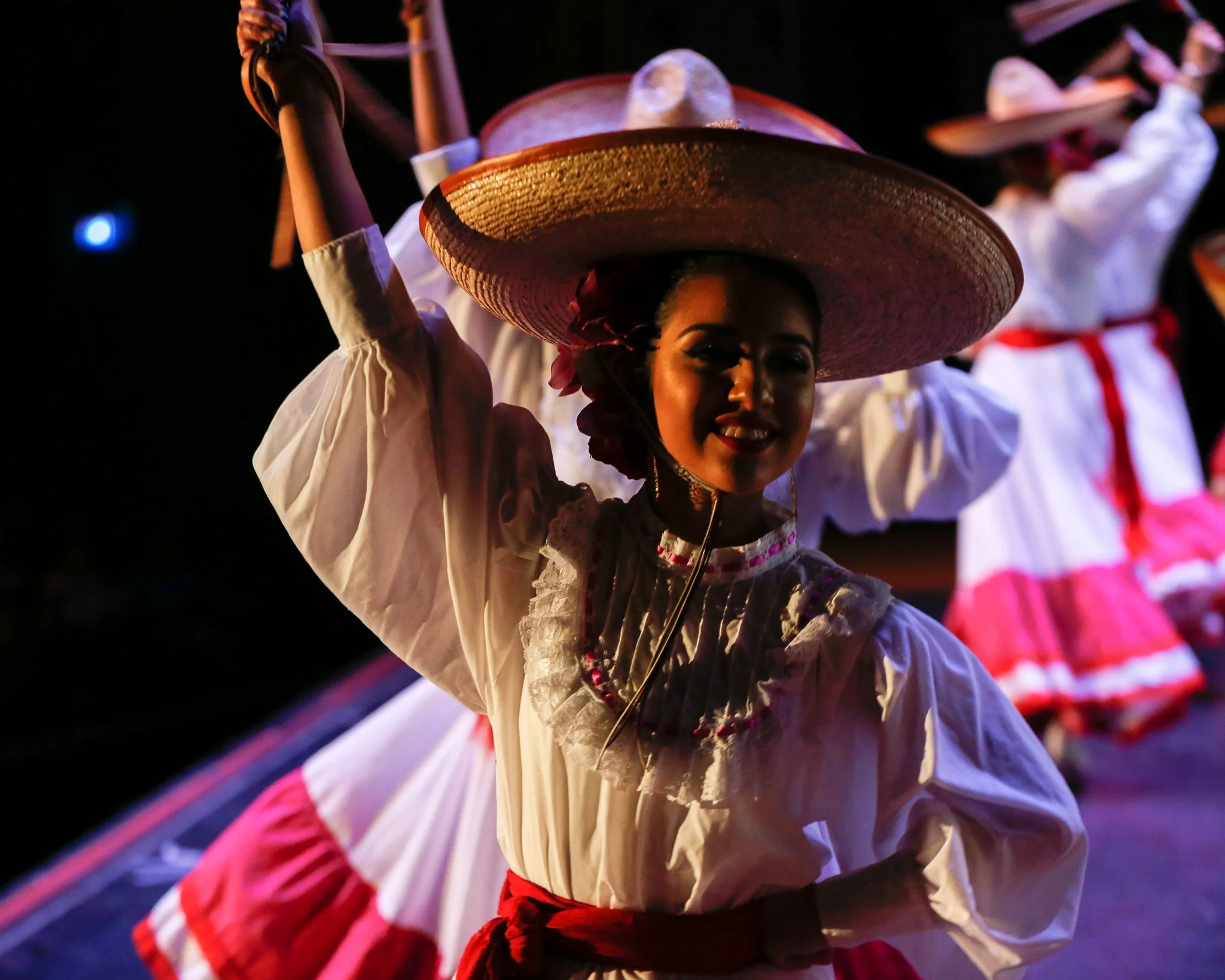 Group of women dressed in traditional Mexican attire, wearing large sombreros and colorful skirts, performing a folkloric dance on stage at night, with stage lights illuminating them.