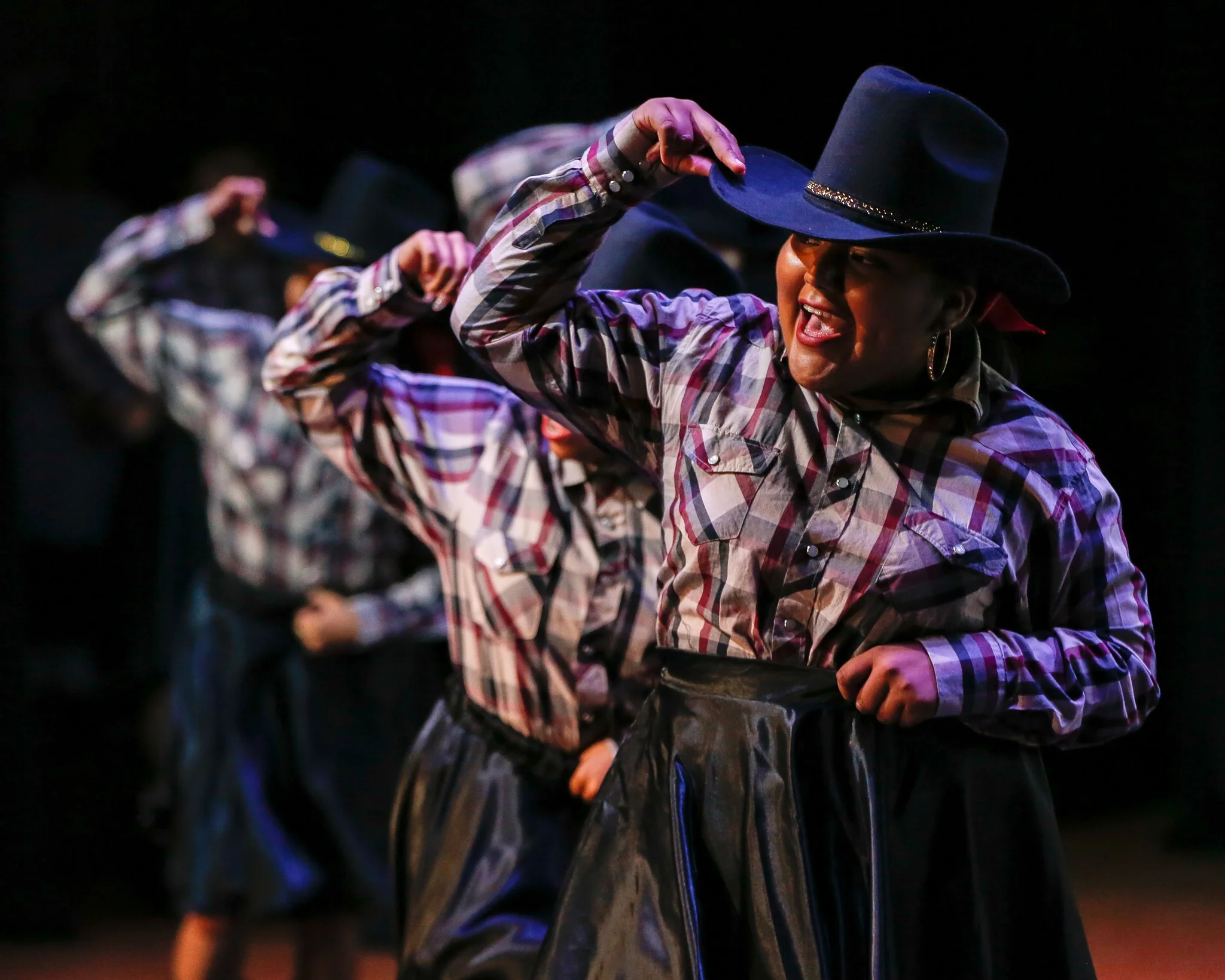 Women dressed in plaid shirts and cowboy hats performing a dance routine on stage.