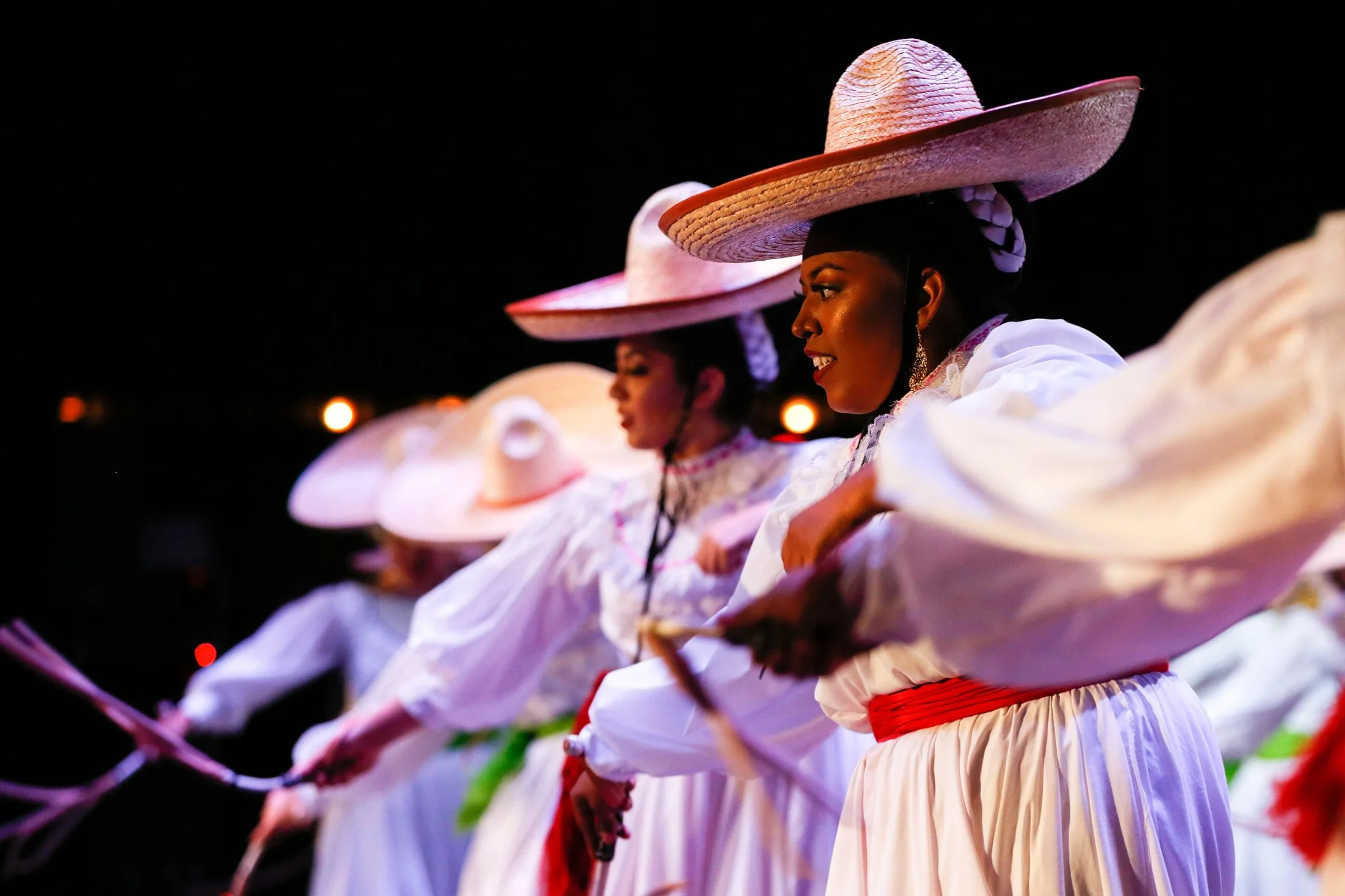 Women dressed in traditional Mexican attire, wearing large sombreros, performing a dance at night.