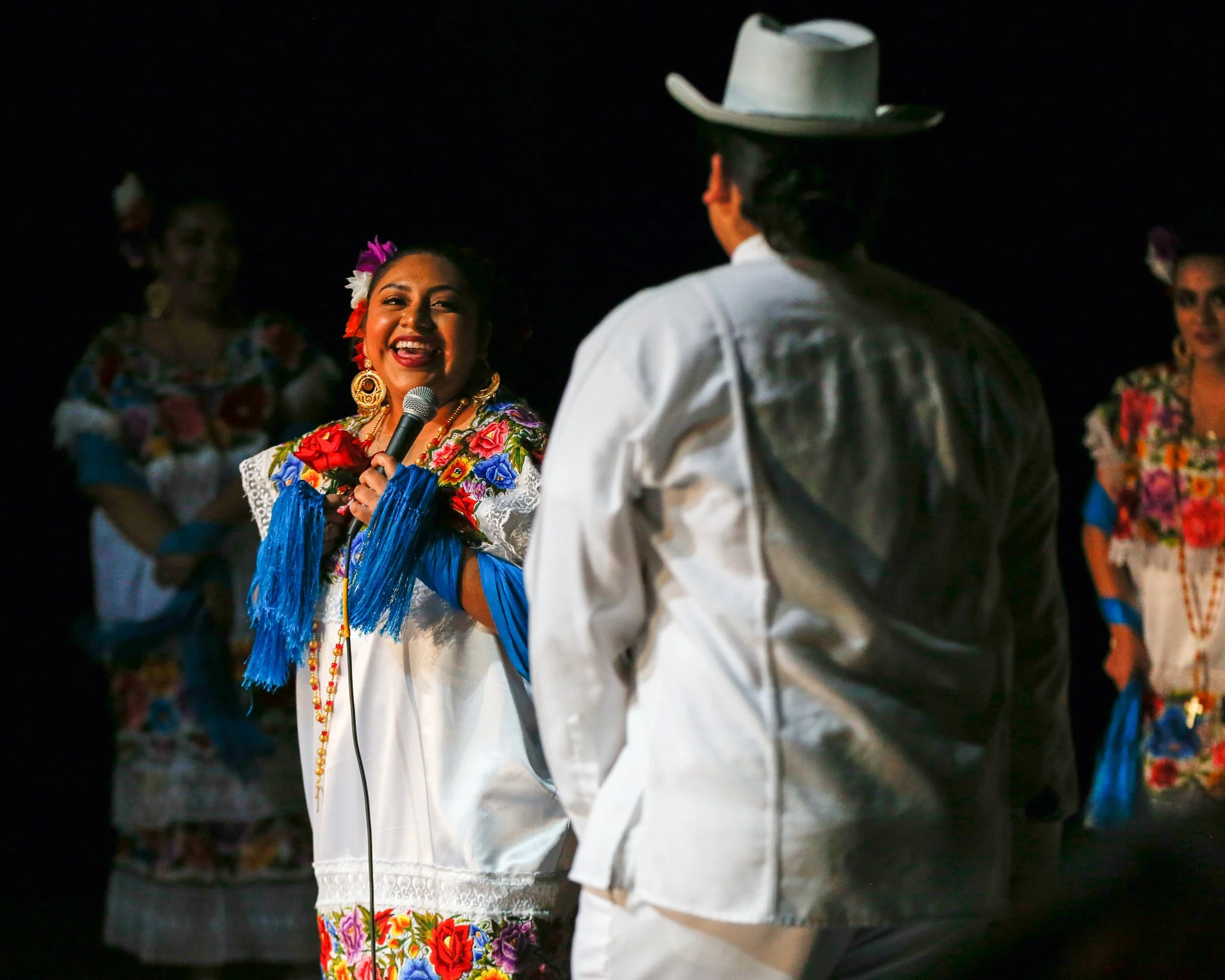 A woman in colorful traditional Mexican dress holding a microphone and smiling at a man wearing a white shirt and gray hat during a cultural performance or celebration, with other women in similar attire in the background.