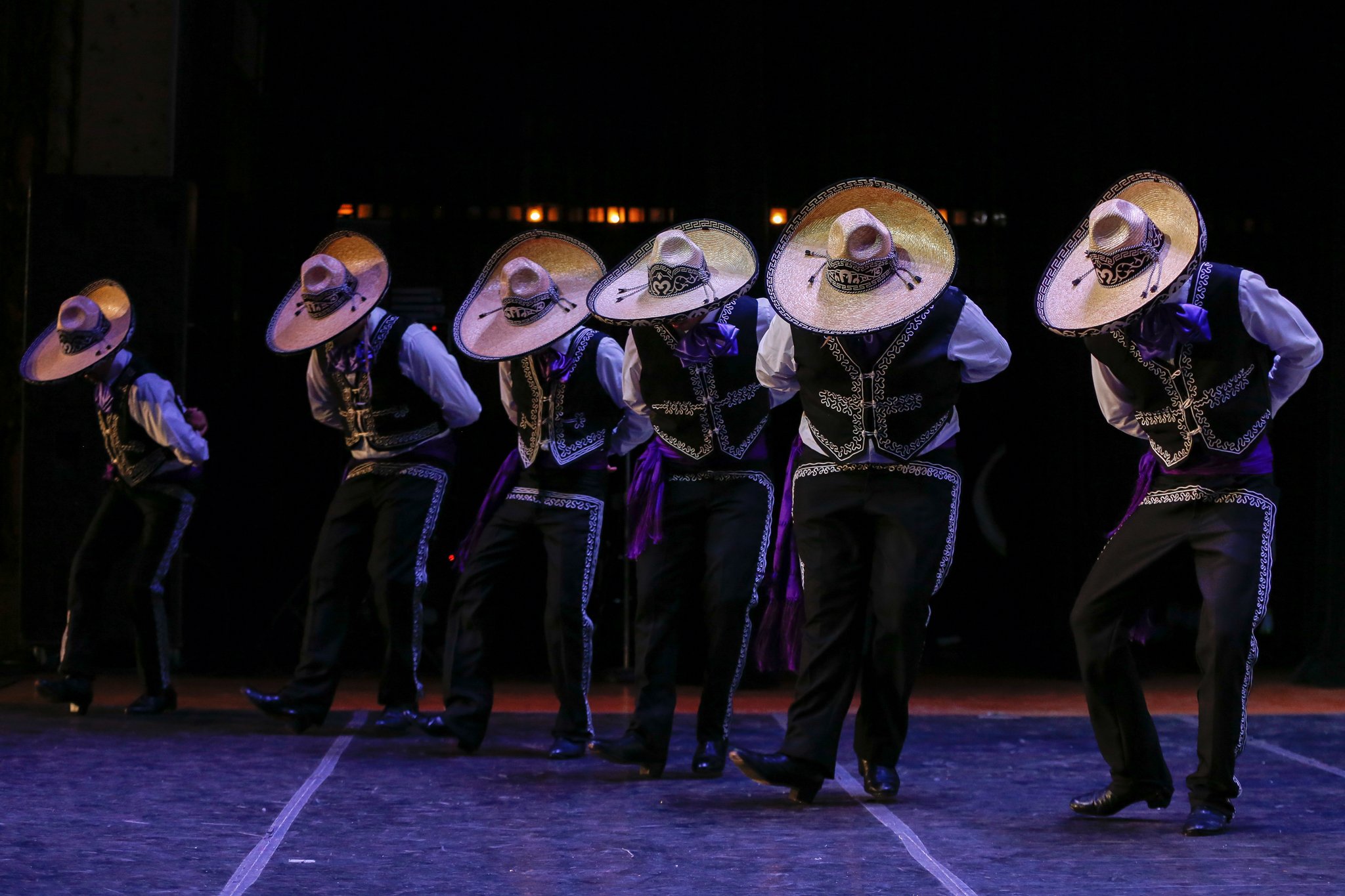 Group of dancers dressed in traditional Mexican attire, performing a folkloric dance on stage under purple lighting, with large straw sombreros and embroidered vests.