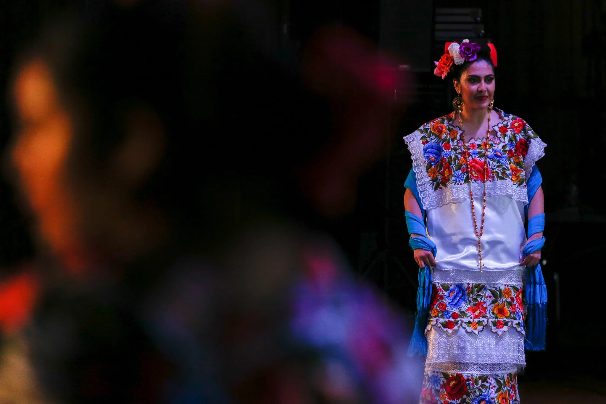 A woman wearing traditional Mexican attire with colorful embroidery, floral accessories, and shawls, stands on a dark stage.