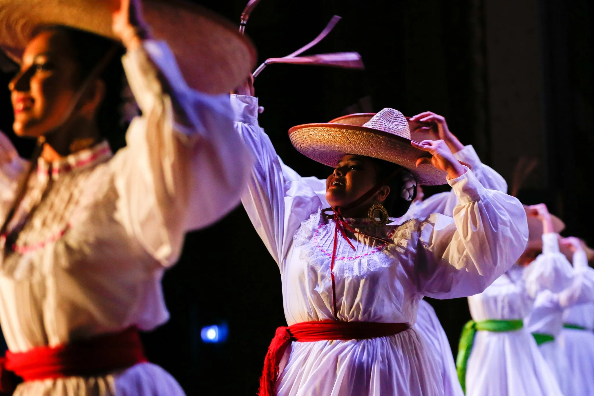 Mexican women wearing white traditional dresses with colorful belts and large sombreros, performing a folkloric dance on stage.