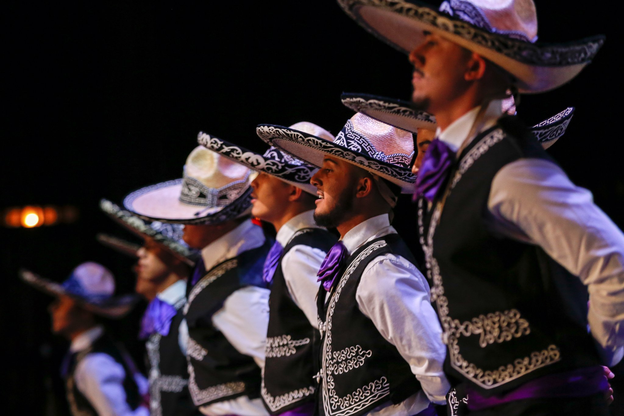 Group of Mexican folk dancers wearing traditional charro attire, including large sombreros, black vests with silver embroidery, and purple neckties, performing during a night event.