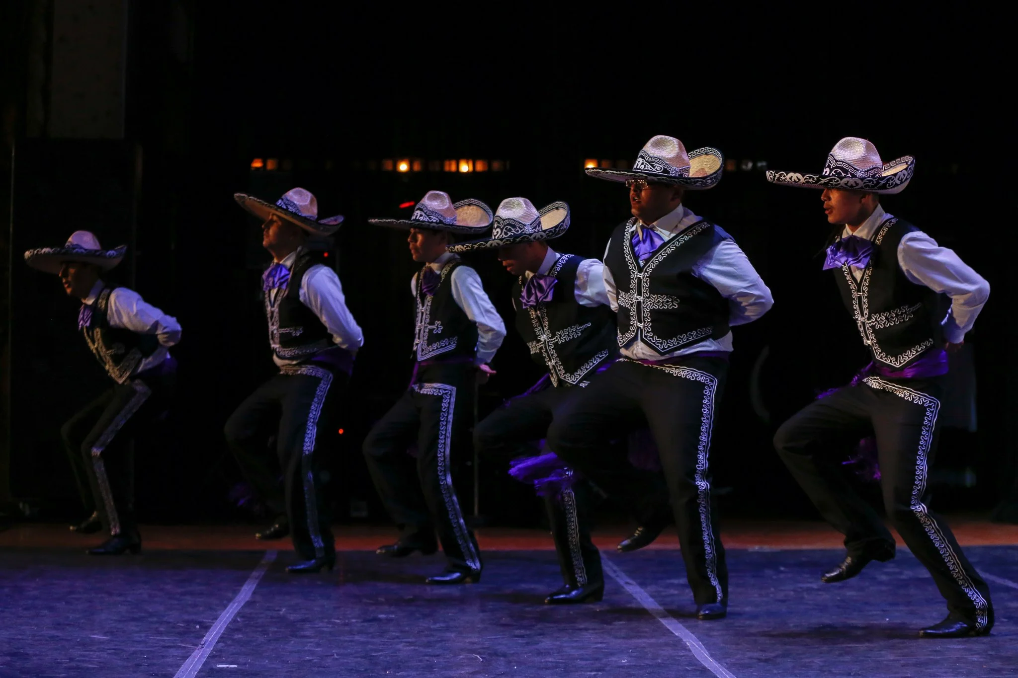 Group of dancers wearing traditional Mexican charro costumes and sombreros, performing a dance on stage in low lighting.
