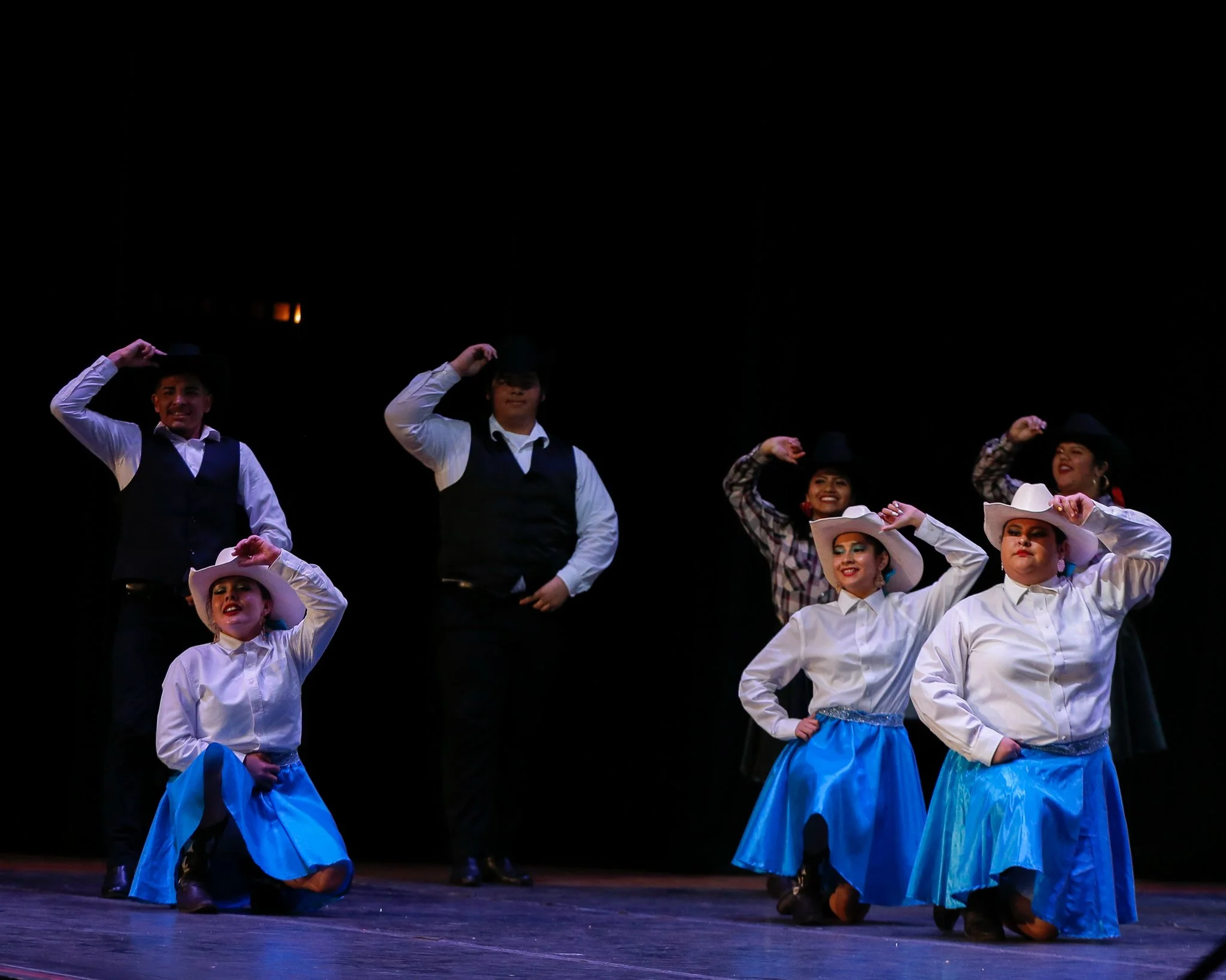 Six dancers performing on stage in Western-style costumes with blue skirts, white shirts, and hats, smiling under stage lights.