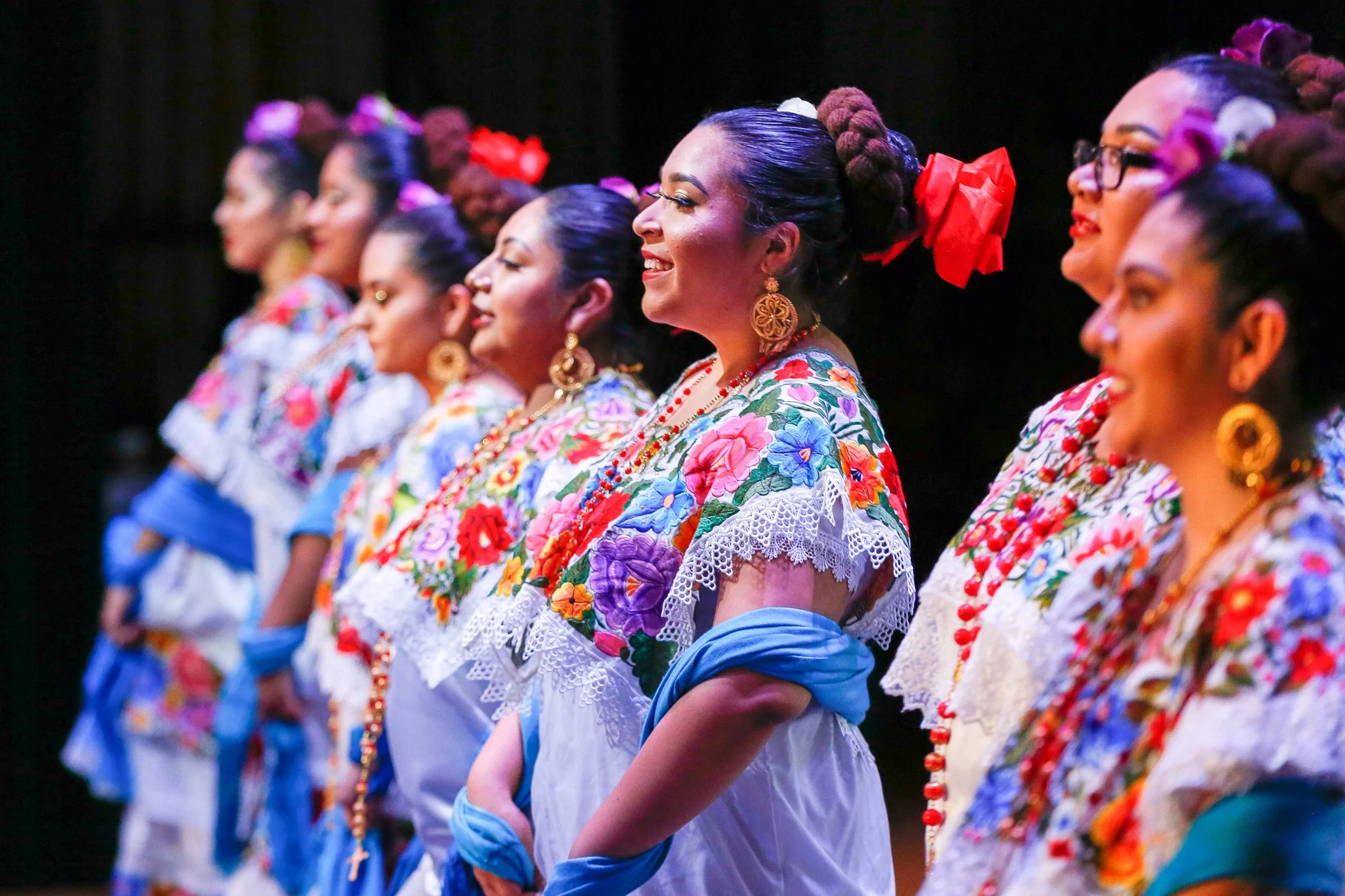 A group of women dressed in traditional Mexican folk costumes with vibrant floral embroidery, colorful sashes, large earrings, and hair styled in braids with ribbons, participating in a cultural dance or celebration.