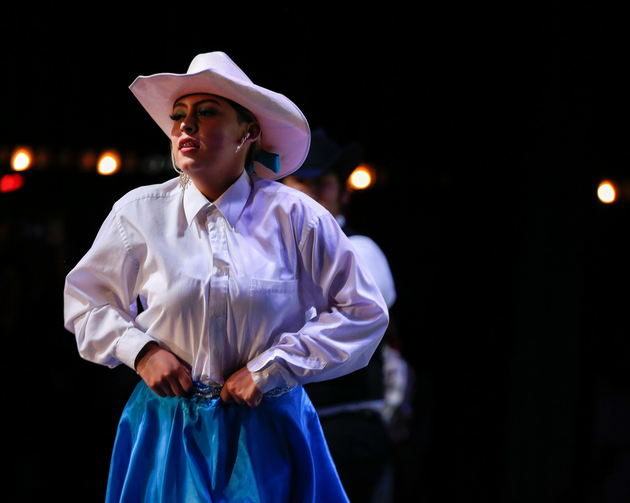 A woman wearing a white cowboy hat, white button-up shirt, and blue skirt stands in a dark setting with small lights in the background.
