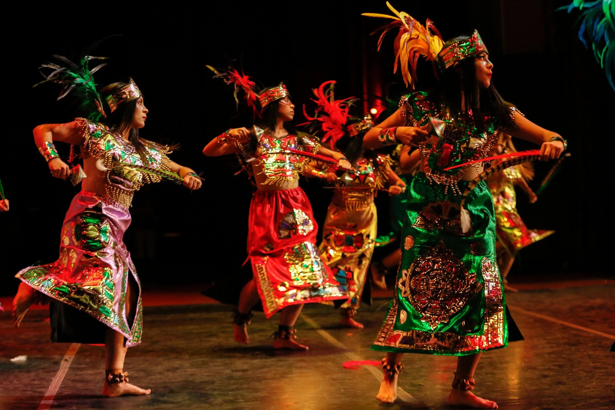 Group of performers in elaborate, colorful costumes with feathers, dancing on stage.