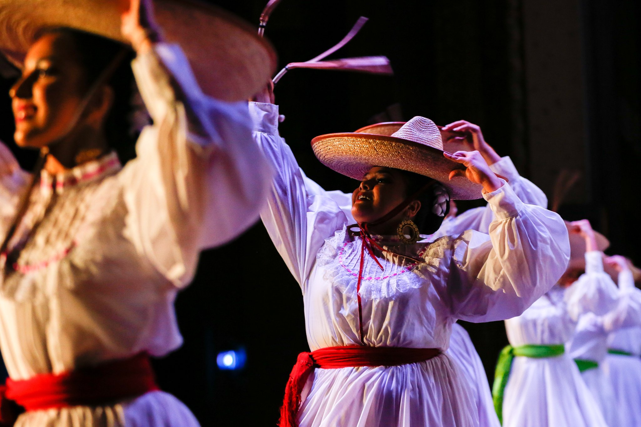 Women dressed in traditional white dresses with colored sashes, wearing large sombreros, perform a dance on stage.
